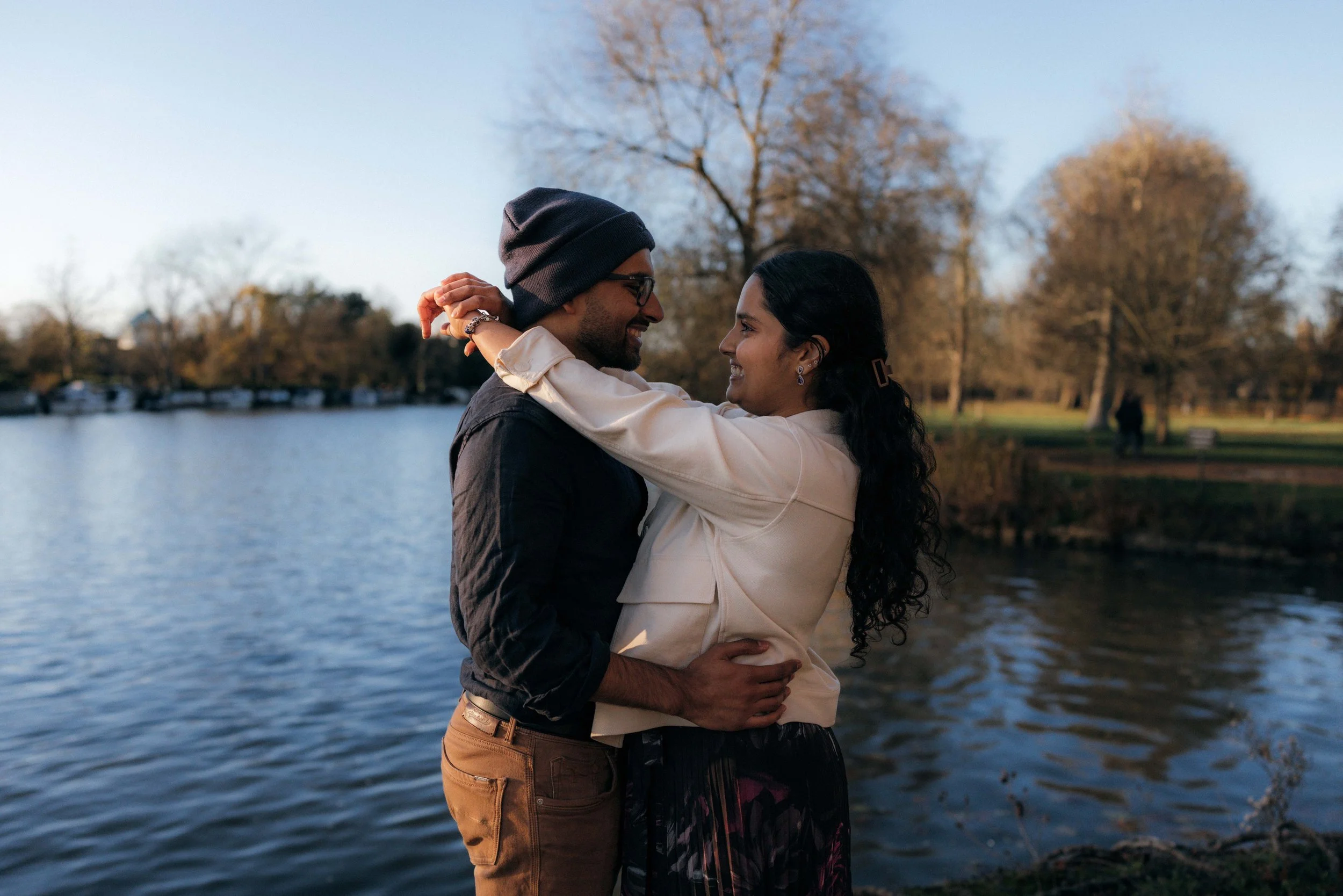 A happy couple embraces by a lakeside during sunset, surrounded by trees with autumn foliage.