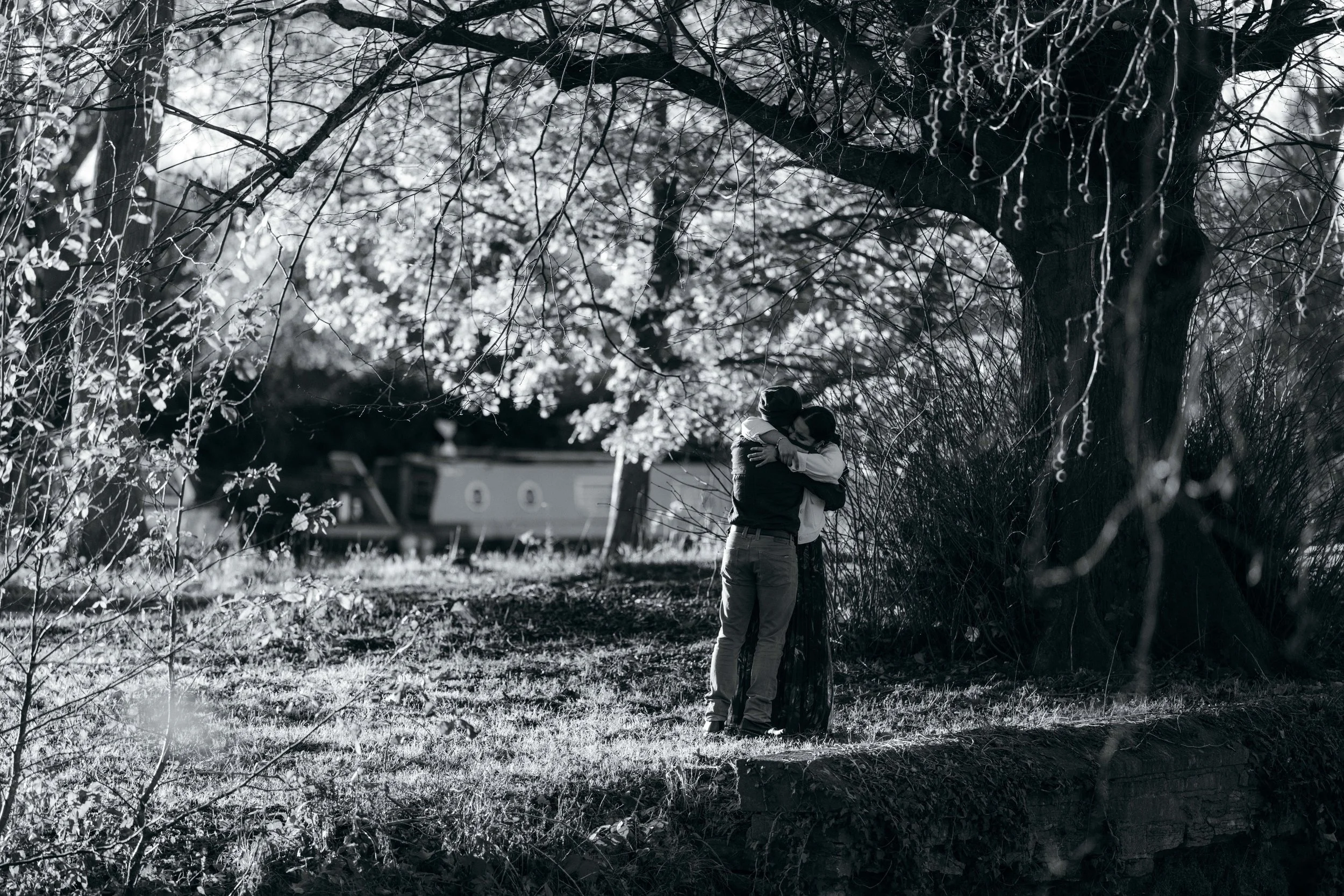 A couple embracing under a large tree with sparse leaves on a grassy area, with a boat visible in the background, in black and white.