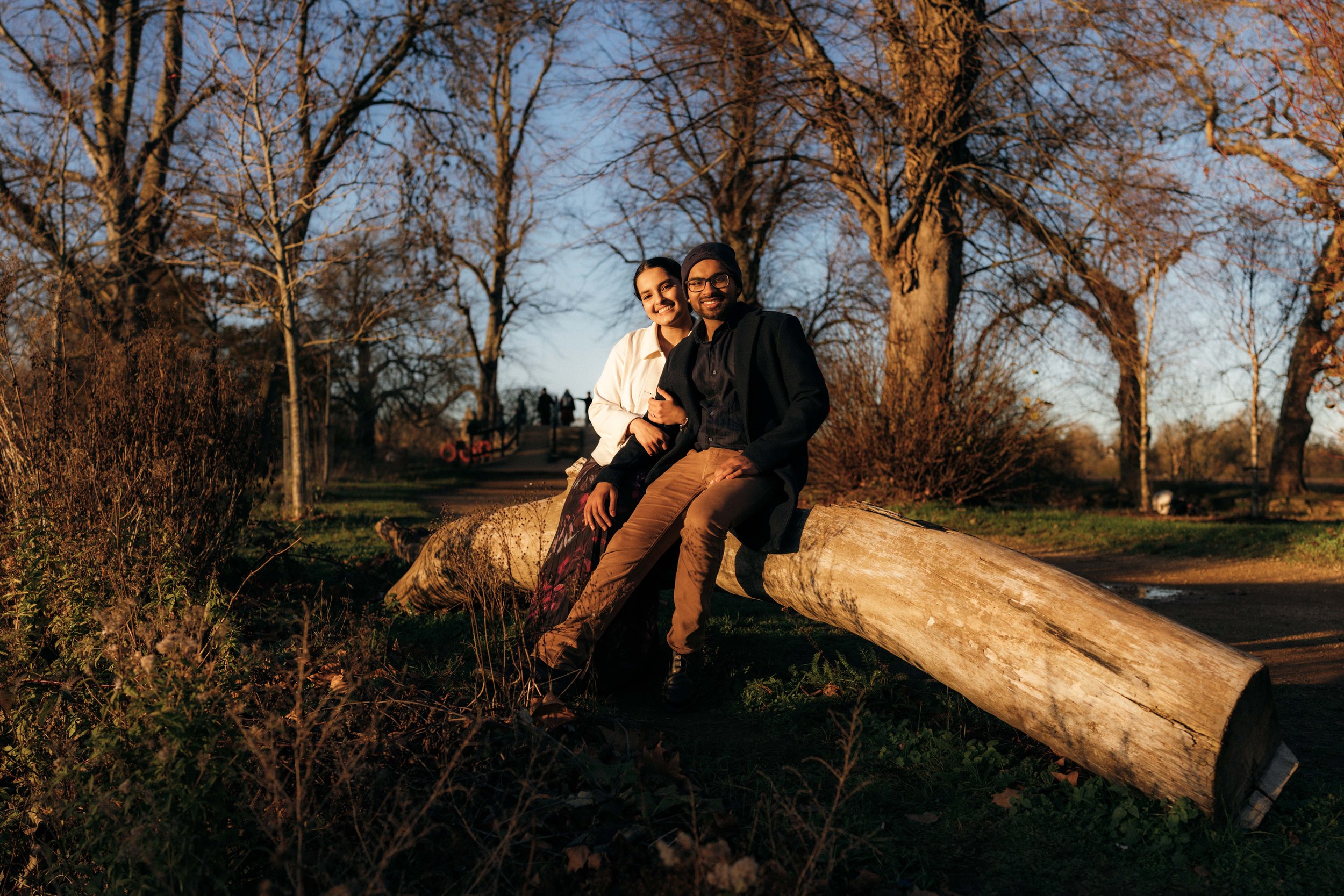 A man and woman sitting on a fallen tree in a park during late afternoon or early evening.