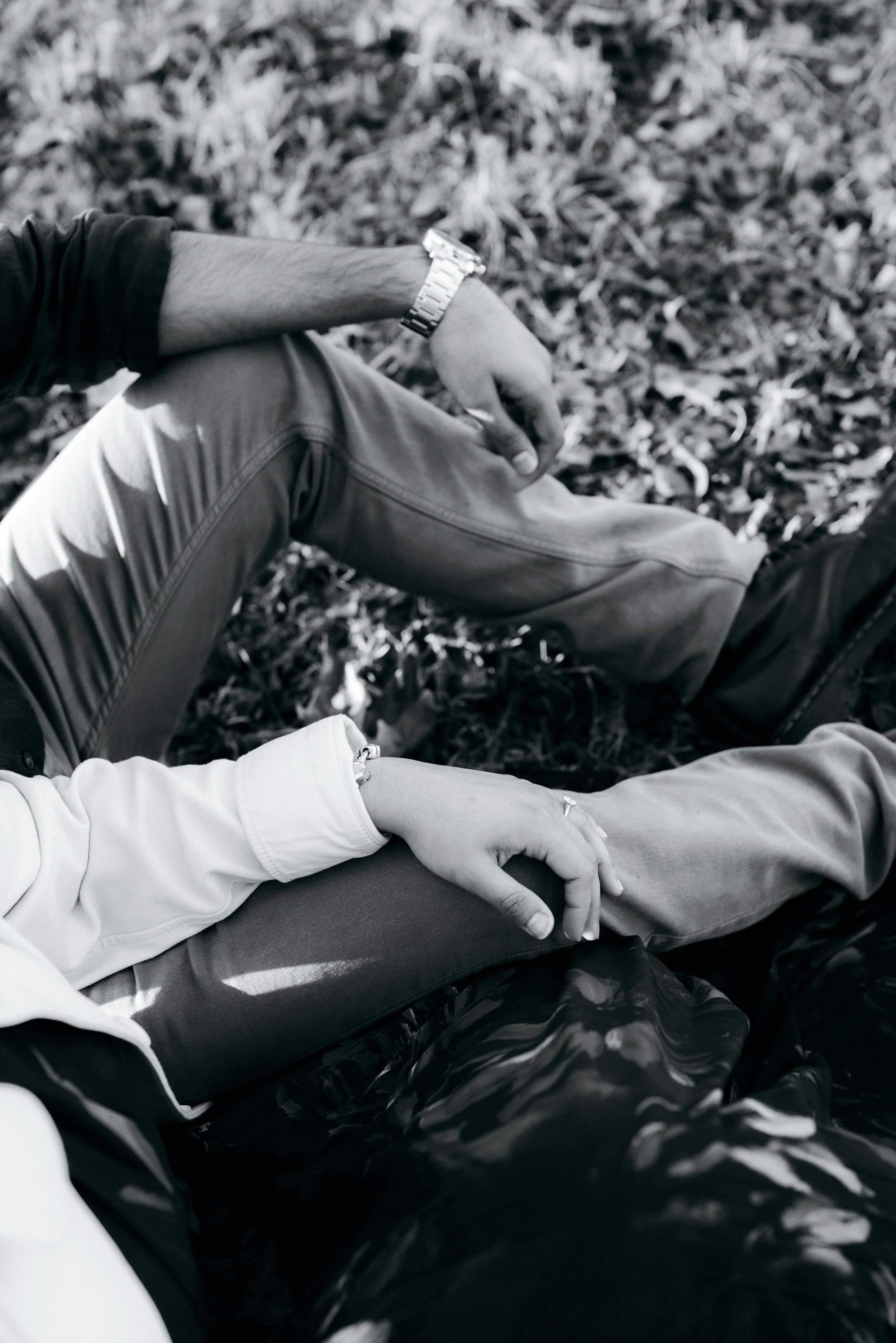 Two people sitting on the ground holding hands, with leaves and grass beneath them. One person wears a watch and light-colored clothing, the other has darker clothing.