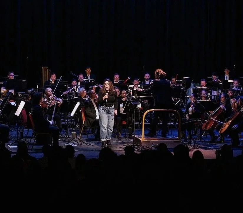 A woman singing with a microphone on stage in front of an orchestra, conductor, and audience.
