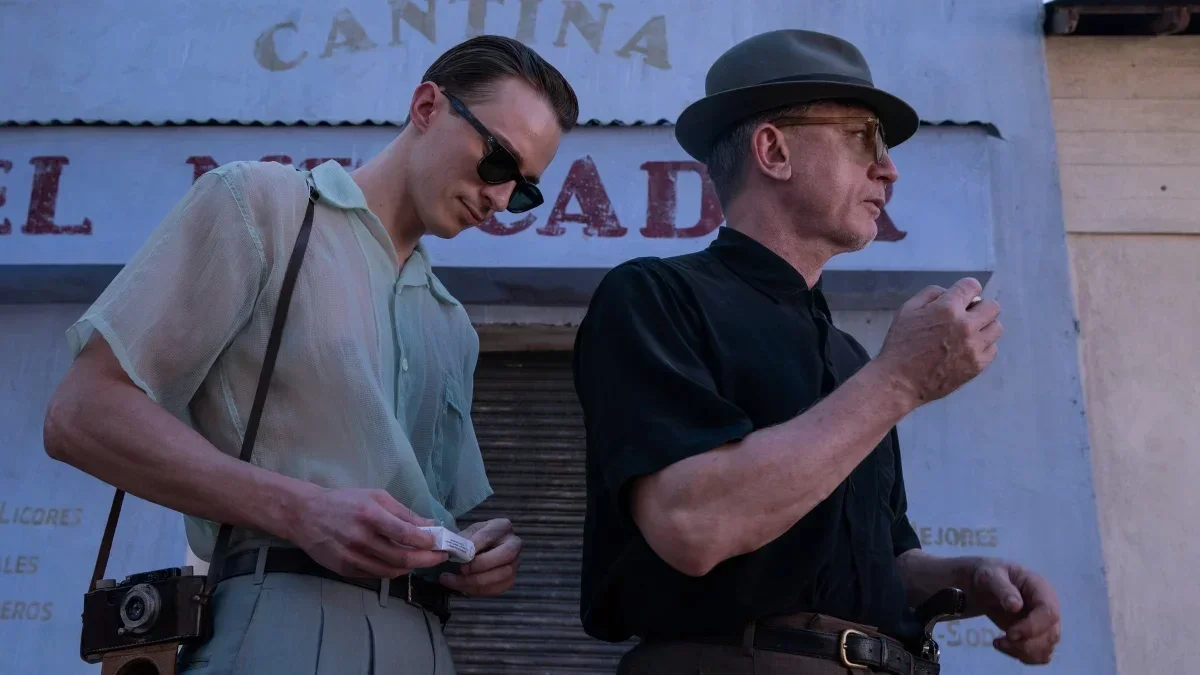 Two men standing outdoors, one looking at a small object in his hands, the other smoking a cigarette. The man on the left wears sunglasses and a loose short-sleeved shirt, with a camera hanging from his neck. The man on the right wears a hat and a dark short-sleeved shirt.