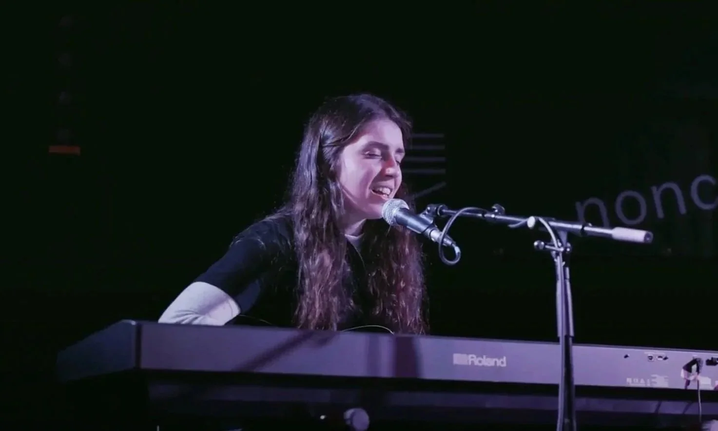 A young woman singing into a microphone while playing a Roland keyboard on a dark stage.