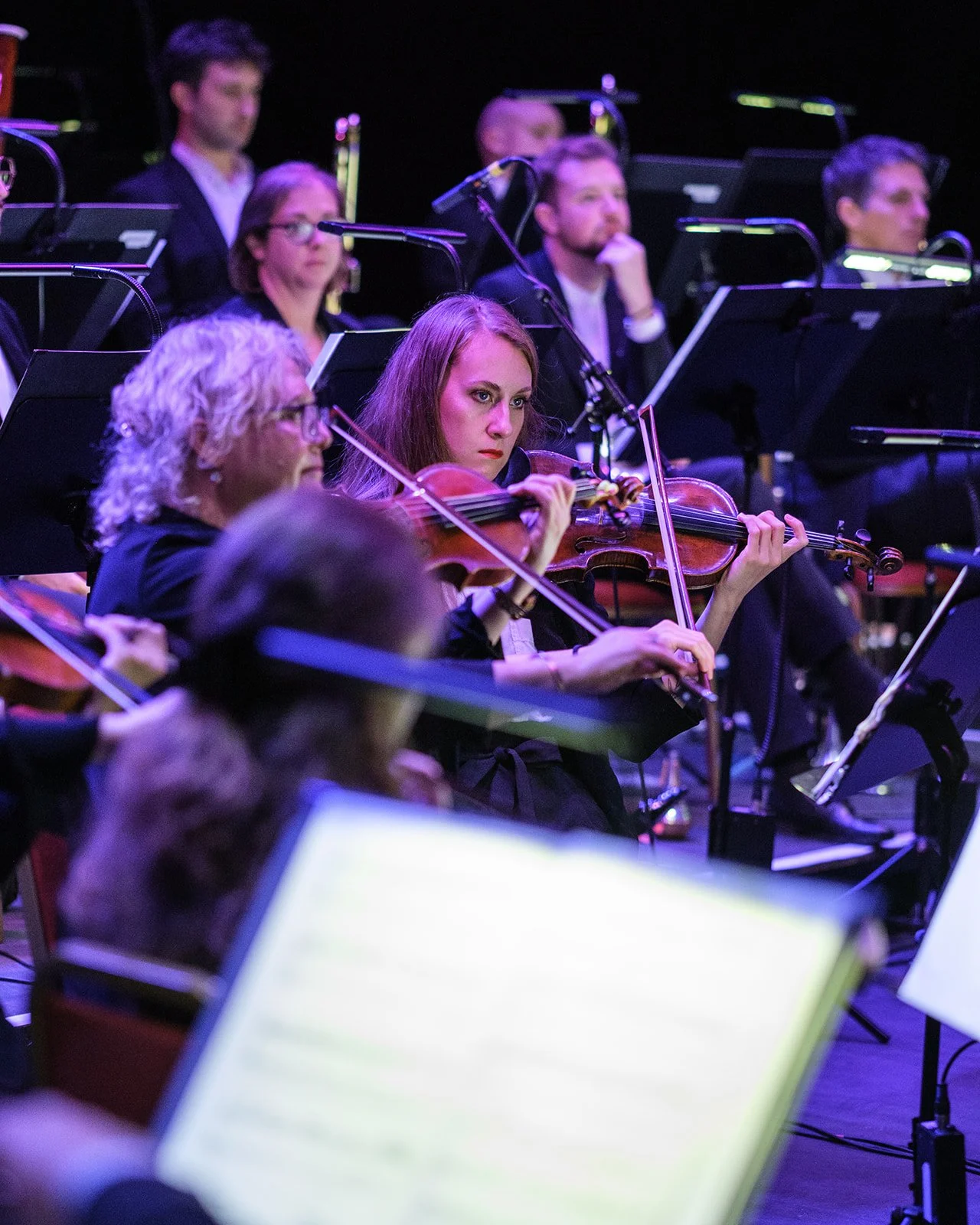 Orchestra performers, including a woman with red hair playing the violin, in a concert setting with music stands and sheet music.