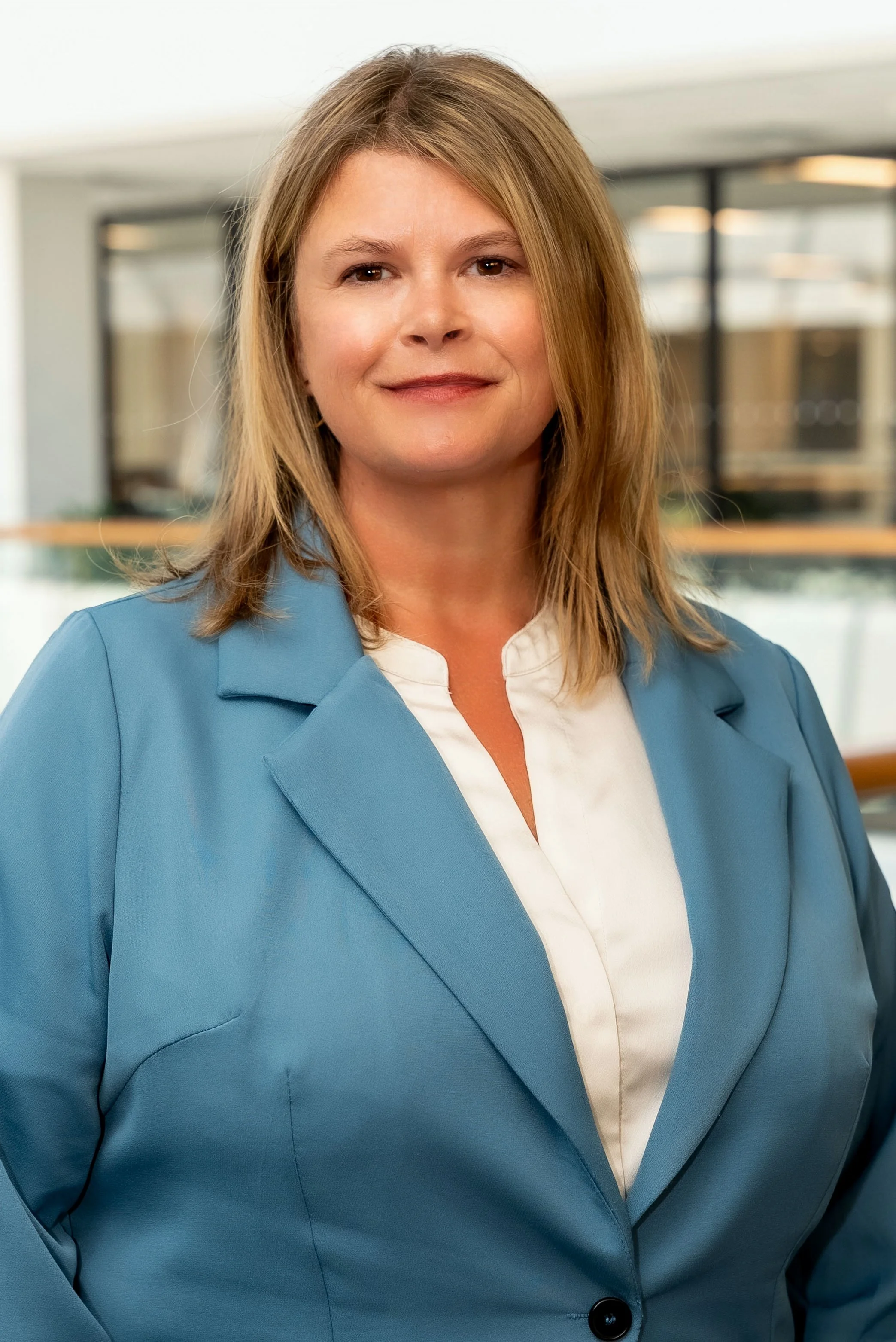 A woman with shoulder-length blonde wavy hair, wearing a navy blazer and a white blouse, standing in an indoor setting with a blurred background.