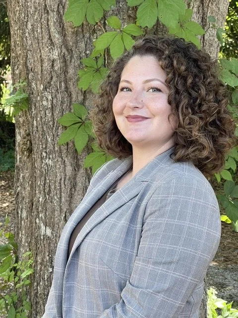 Woman with short curly hair standing outdoors in front of a green bush, wearing a gray button-up shirt, beige pants, a black belt, and a smartwatch, smiling at the camera.