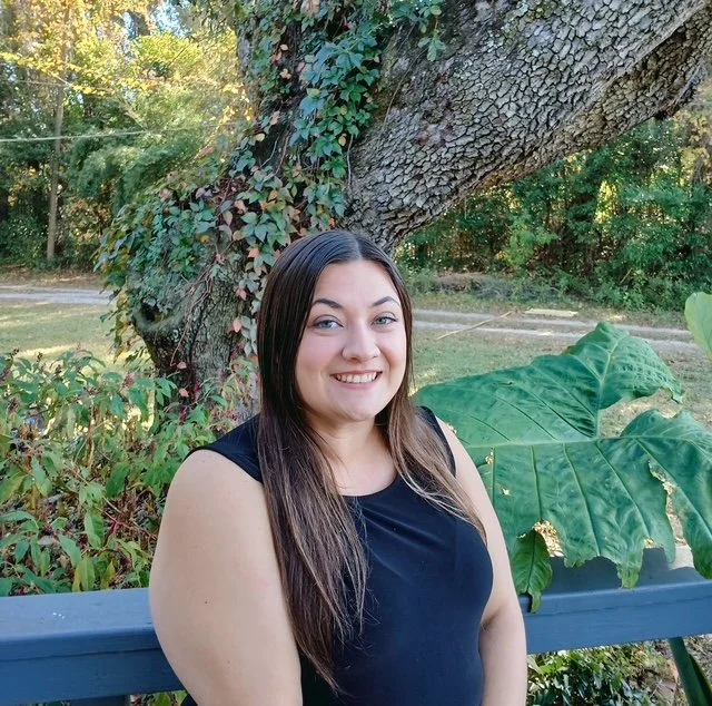 A smiling woman with long brown hair, wearing a black blazer and white top, standing against a light gray background.