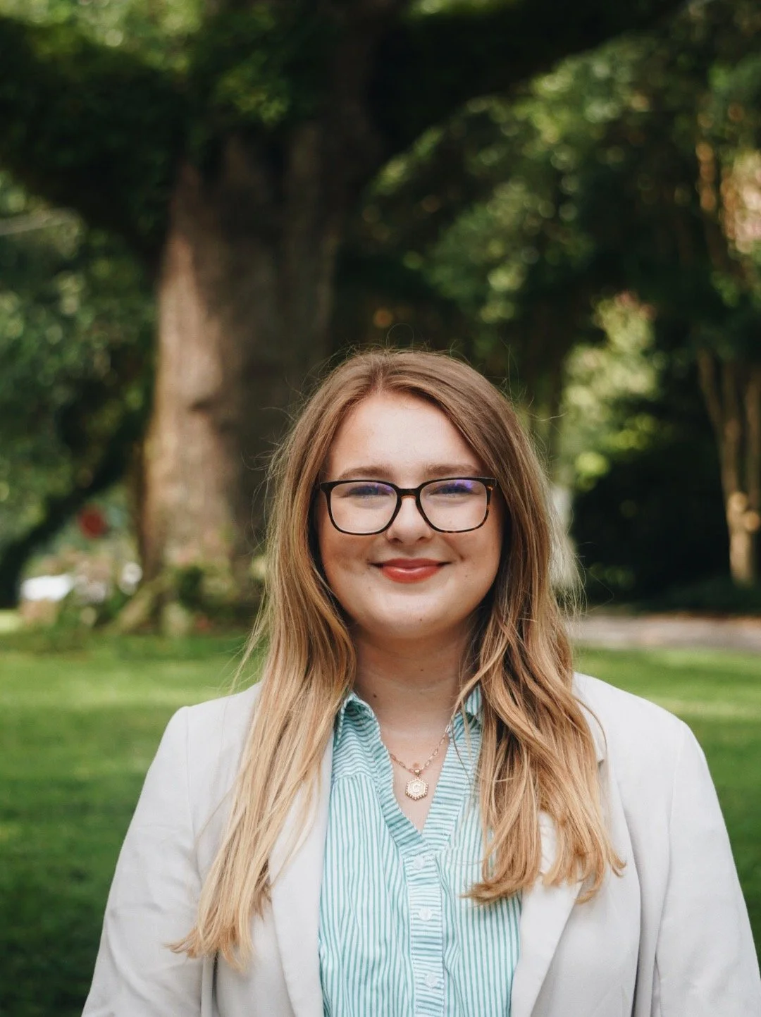 A smiling woman with long brown hair, wearing a black blazer and white top, standing against a light gray background.
