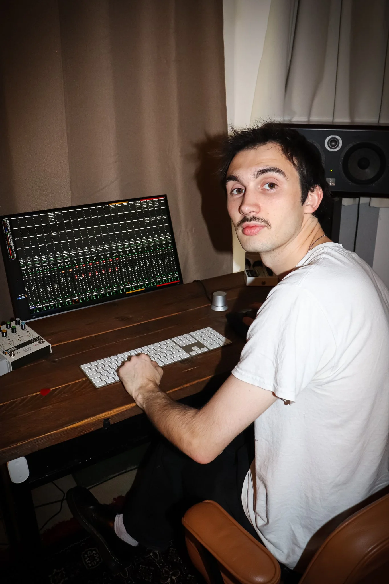 A young man with dark hair and a mustache, wearing a white t-shirt, is sitting at a wooden desk in front of an electronic music mixer and a computer keyboard. There is a large monitor displaying a digital audio mixing interface, and a pair of studio speakers is visible behind him. The setting appears to be a home or professional music studio.