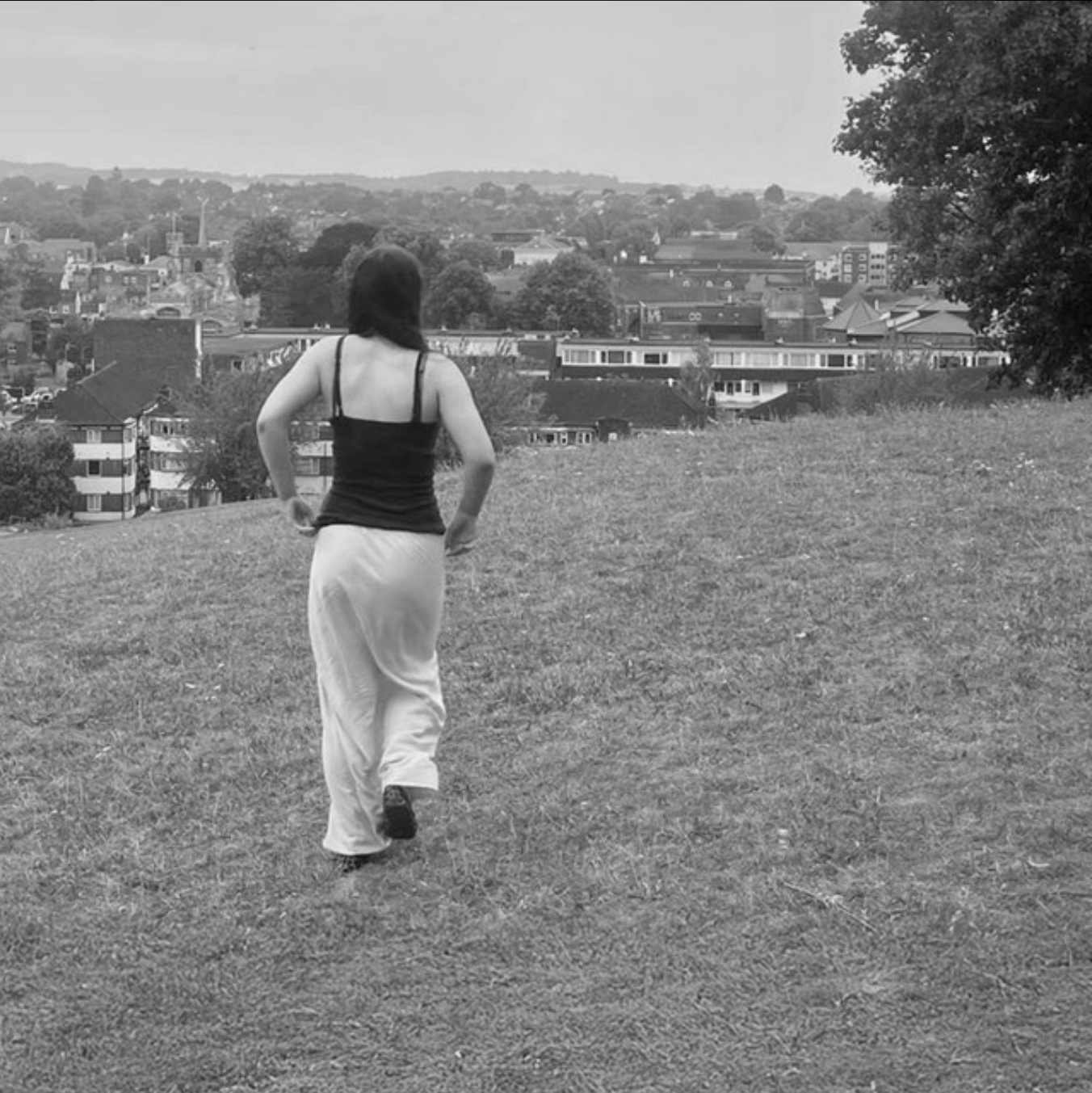 A woman walking away on a grassy hill with a cityscape in the background.
