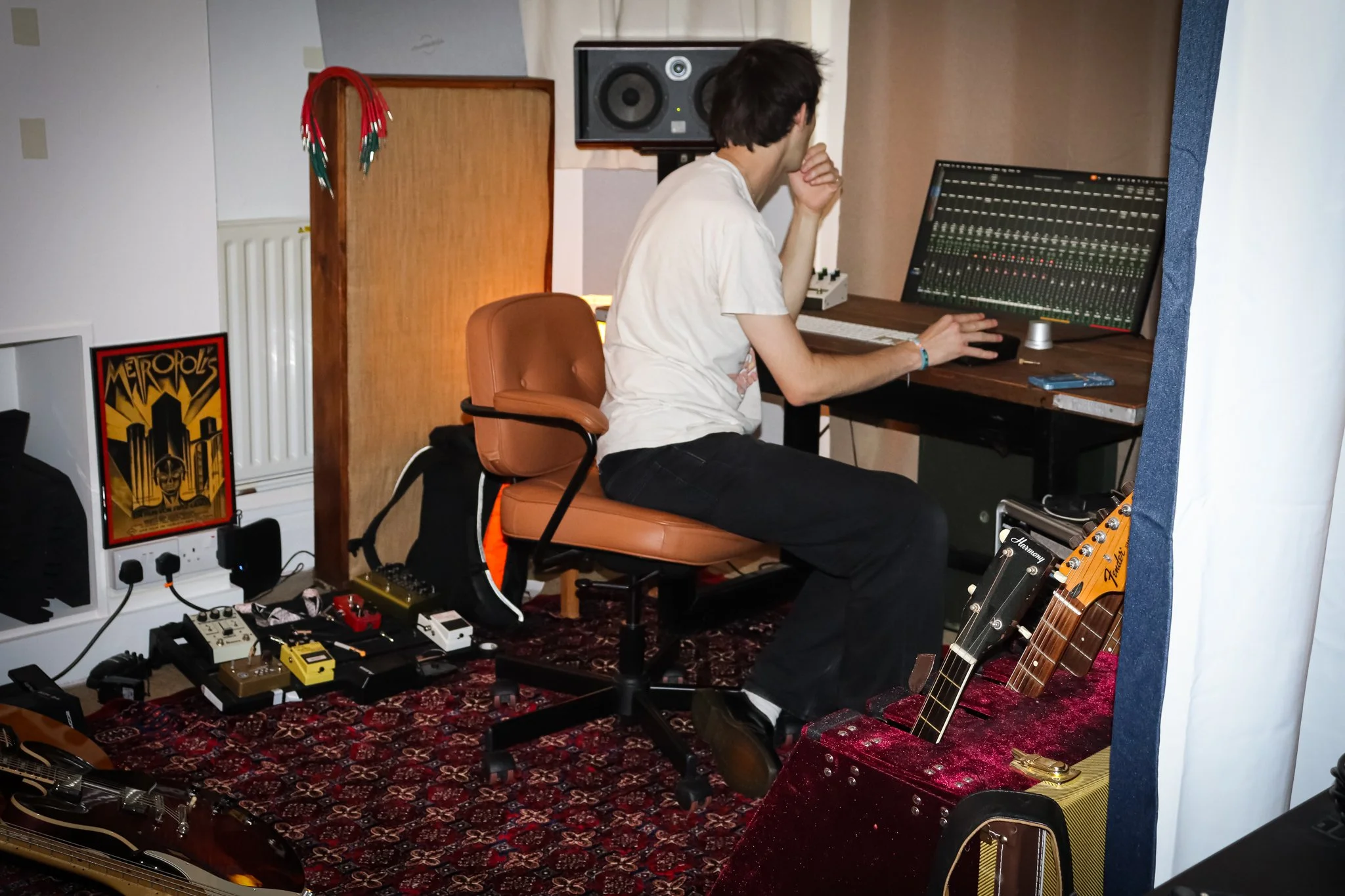 A man with dark hair sits at a wooden desk surrounded by music equipment, including a computer with a digital audio workstation, a keyboard, a guitar, and numerous effect pedals on the floor. The room has posters and a rug, indicating a music studio.