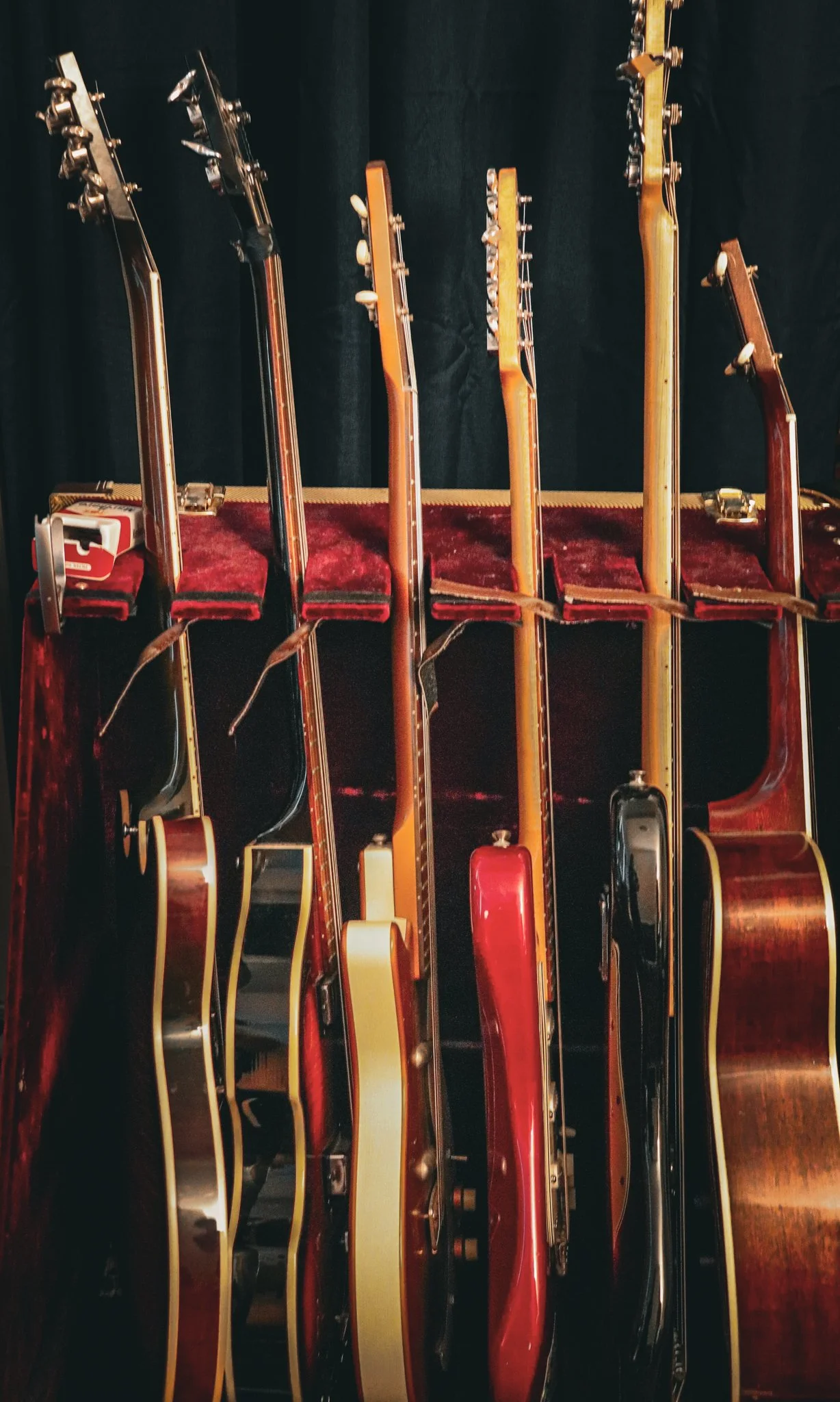 Multiple guitars with different wood finishes and colors leaning against a red velvet-lined surface, against a black background.