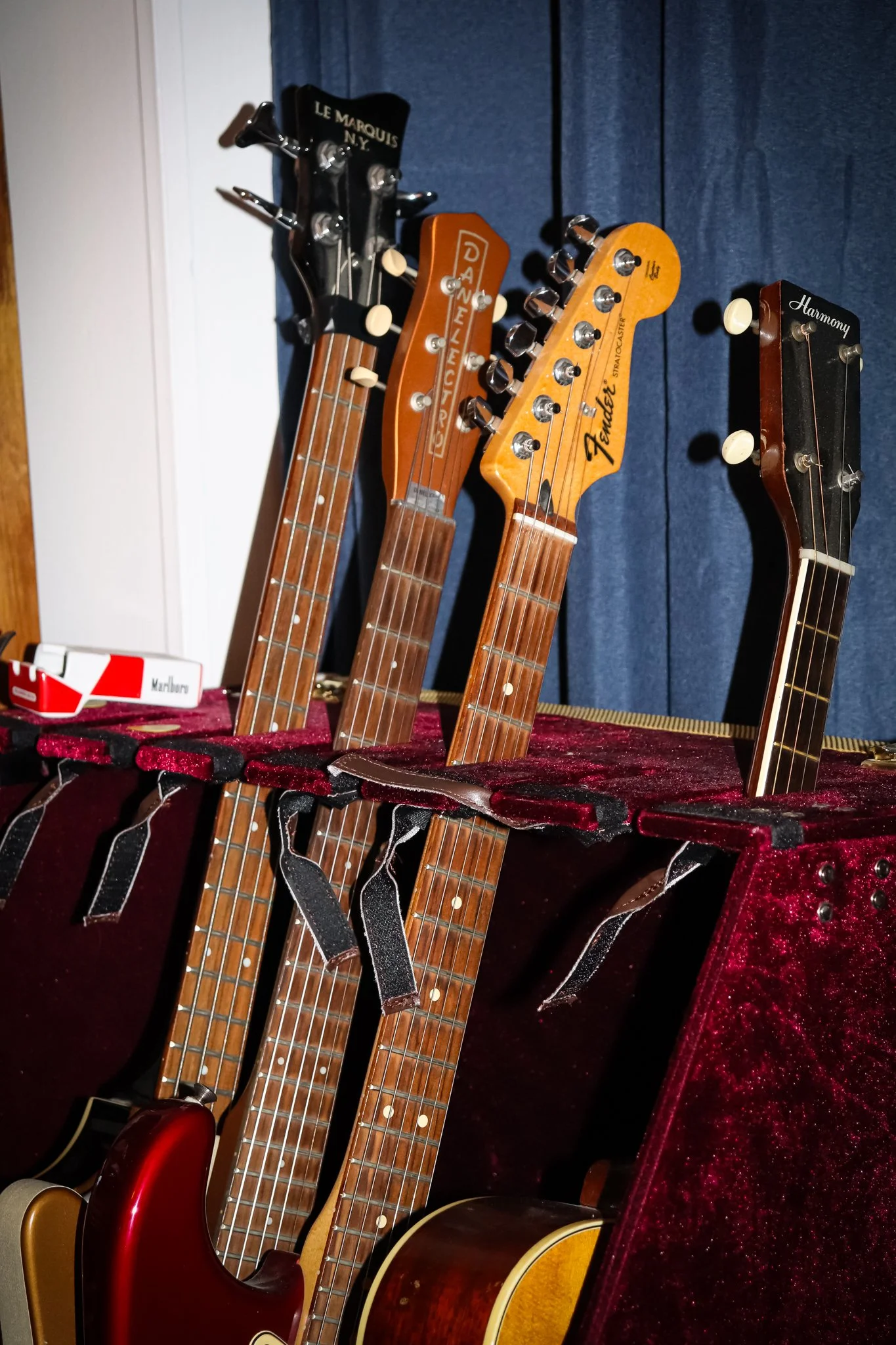 Five guitars in a rack, with the headstocks visible along with tuning pegs and part of the bodies, placed on a red velvet-lined stand, with a box of Marlboro cigarettes visible on the left side.