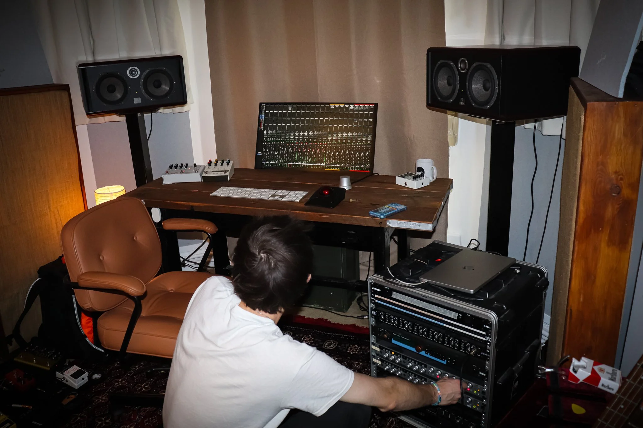 A person with short dark hair wearing a white shirt is working with audio equipment in a home recording studio. The studio has a wooden table with a large audio mixer, computer keyboard, and mouse. Two large black speakers are mounted on stands on either side of the table, and a window with white curtains is behind them. There are various electronic devices and cables around the person, who is seated on the floor in front of a rack of audio processing gear.