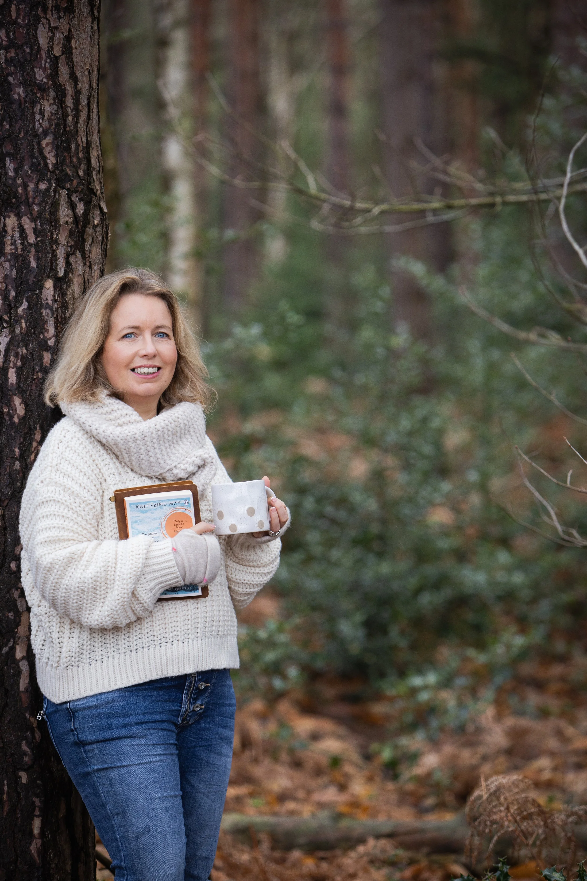 Sarah Robinson standing beside a tree in woodland holding a mug and Katherine May’s book Wintering.