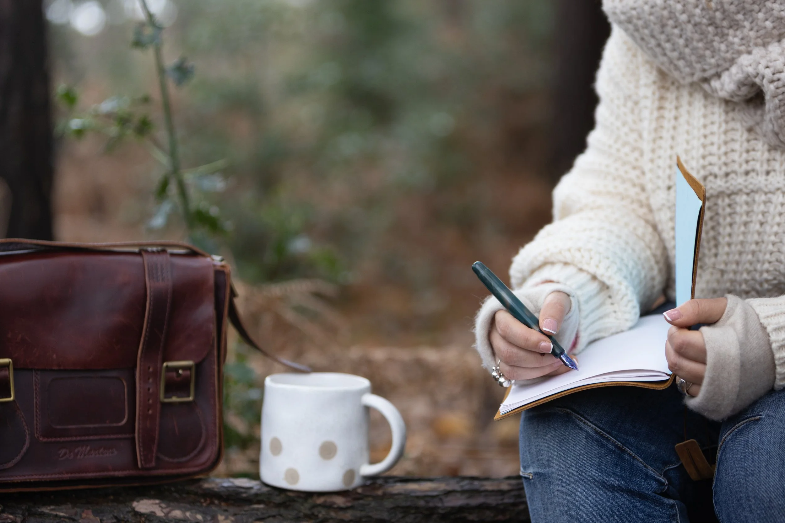 Person sitting outdoors writing in a notebook with a pen, with a brown leather bag and a white mug on a log nearby.