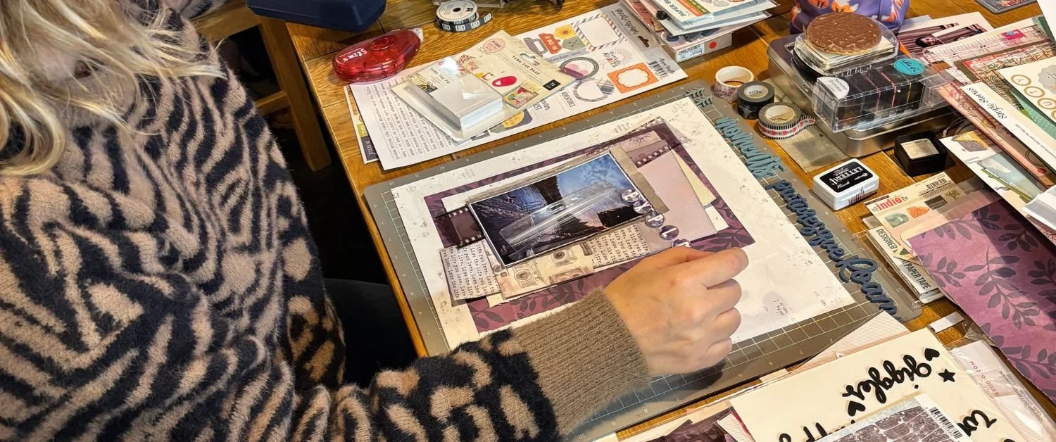 Sarah arranging photos and paper on a scrapbook page at a craft table.