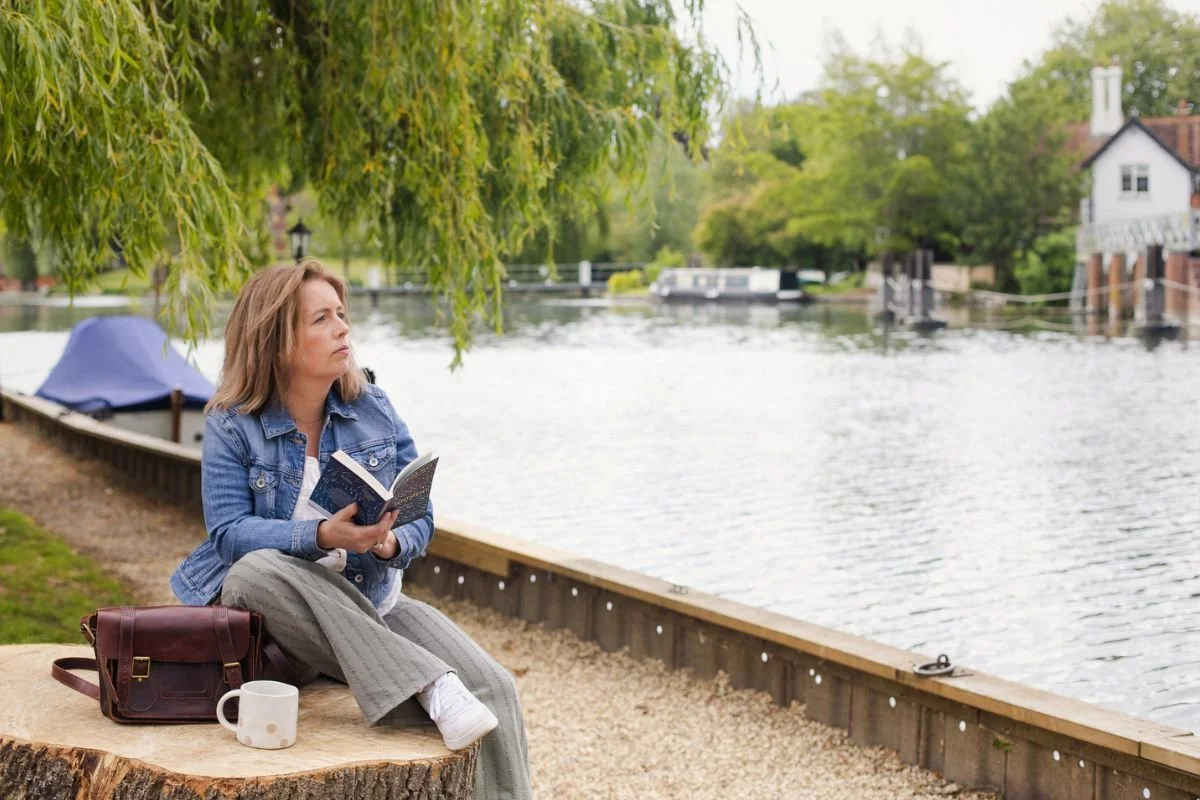 Sarah sitting beside a river with a book and a cup of tea, taking time to slow down and enjoy a quiet moment.