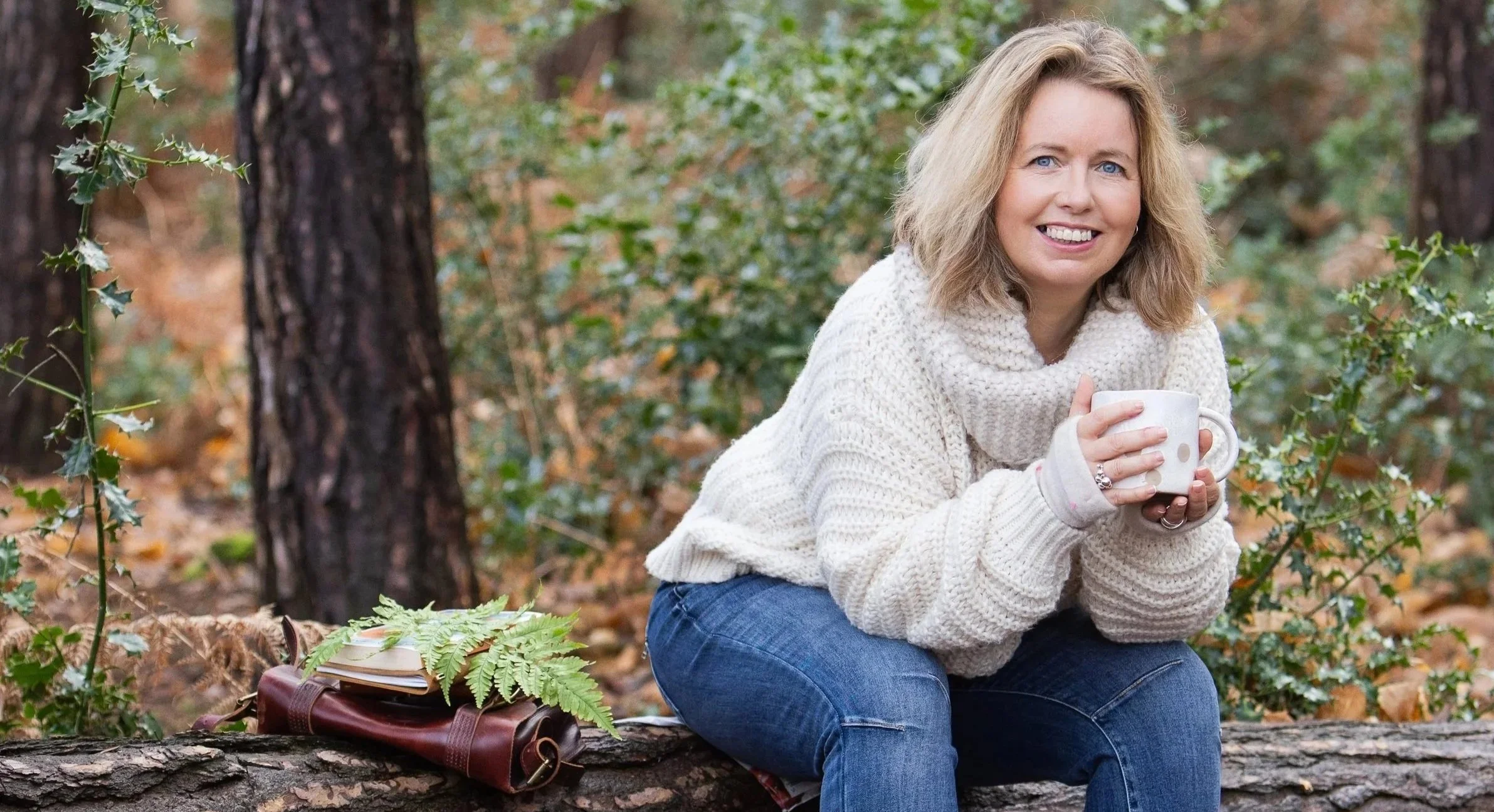 Sarah Robinson sitting on a fallen tree in the forest holding a mug, reflective wellbeing writer and founder of Mindful & Me.