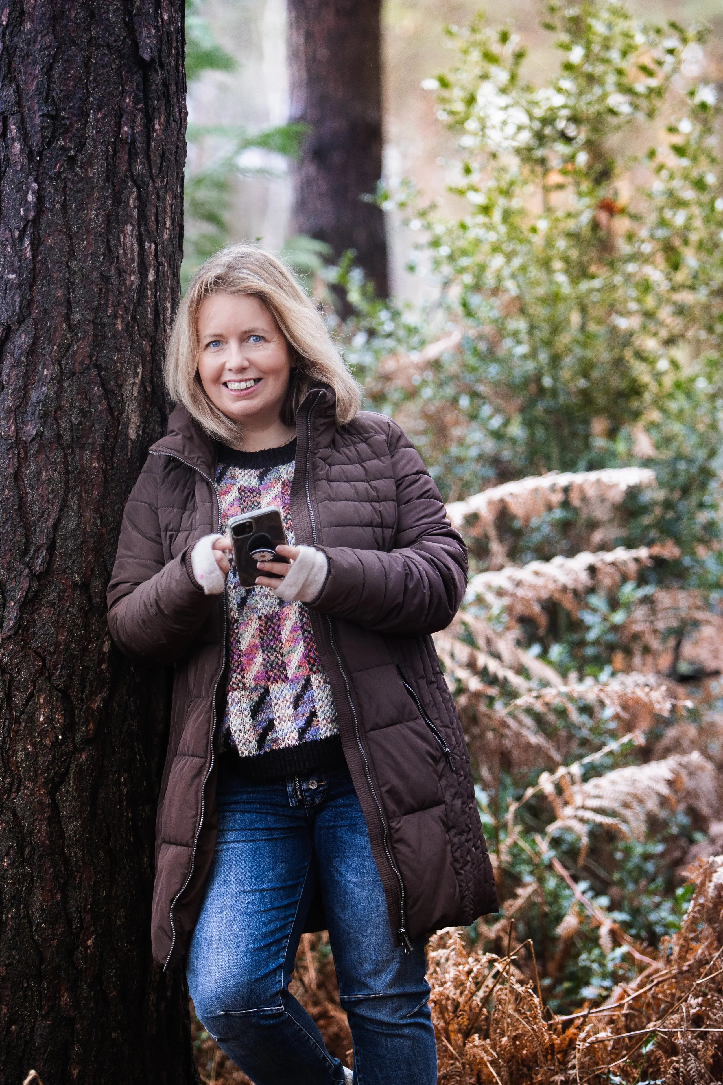 Woman standing by a tree in woodland holding a phone.