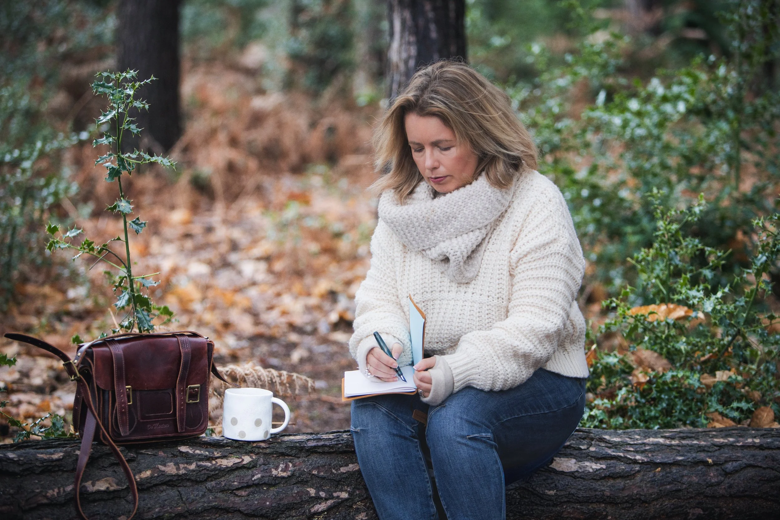 Sarah Robinson writing in a notebook while sitting on a fallen tree in woodland, reflecting outdoors with a mug and bag beside her.