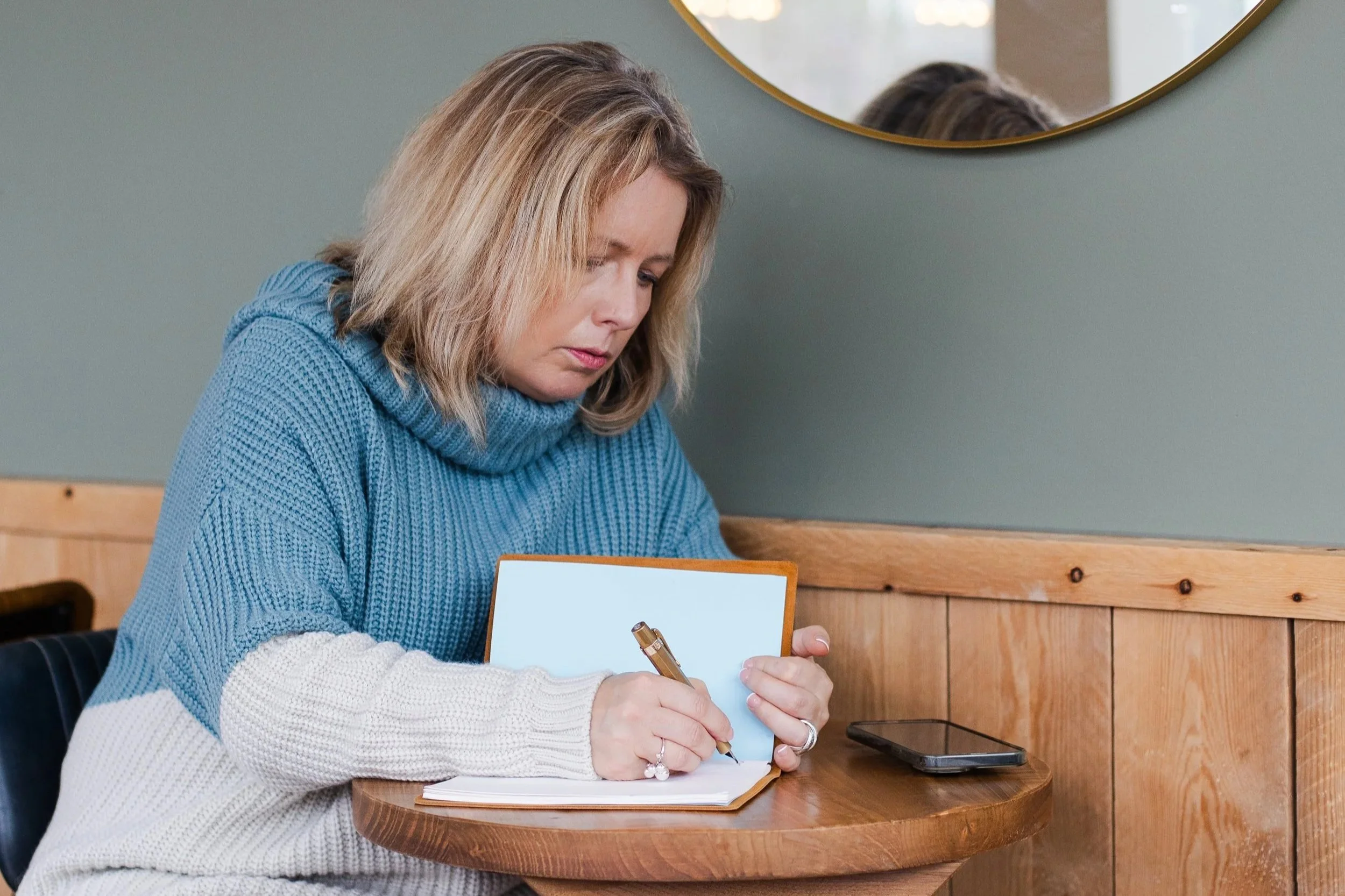 Woman sitting at a café table writing in a notebook with a drink beside her.