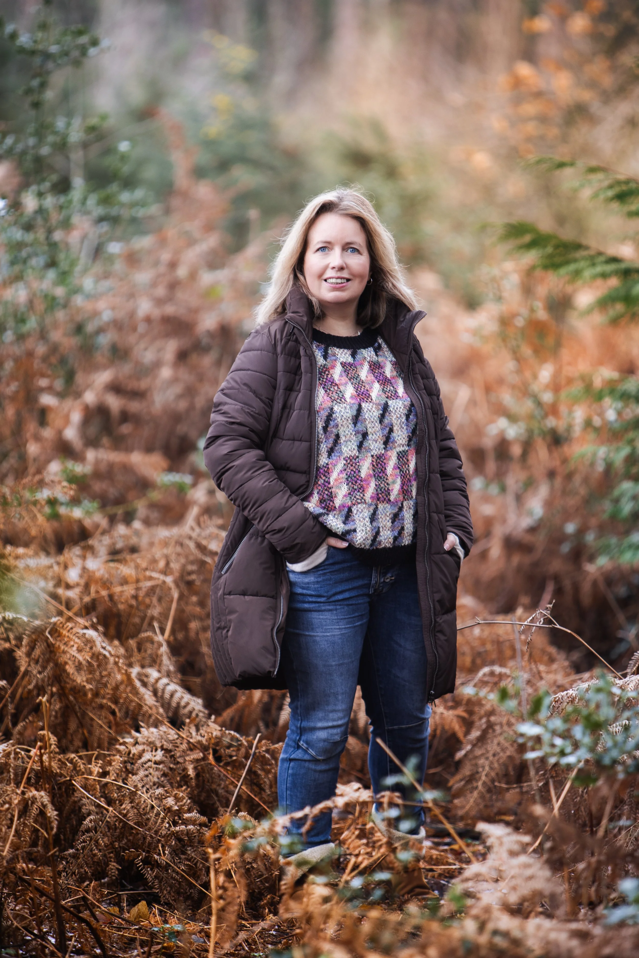 Woman standing in woodland with hands in pockets, looking towards camera.