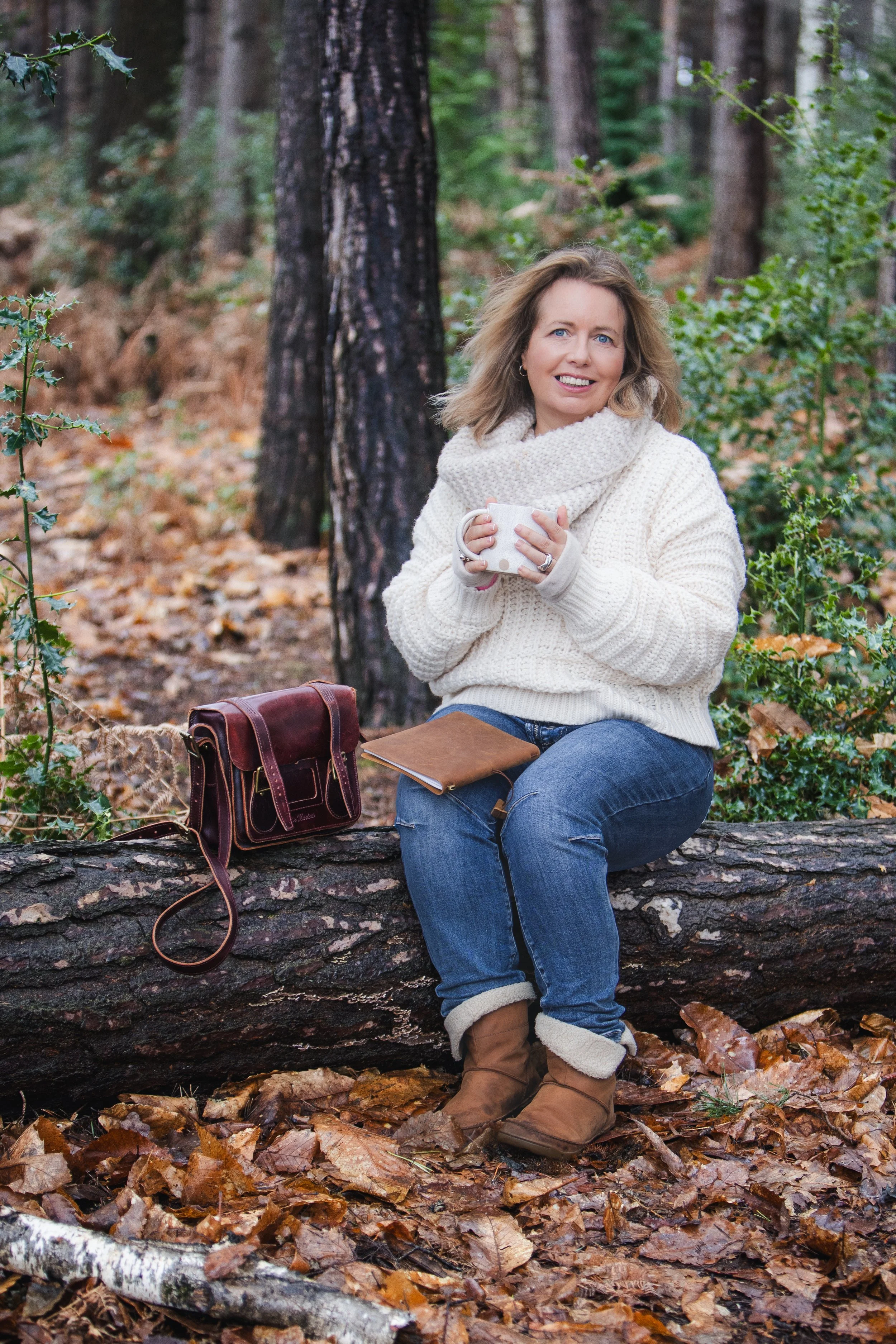 Woman sitting on a log in woodland holding a mug, with notebook and bag beside her.