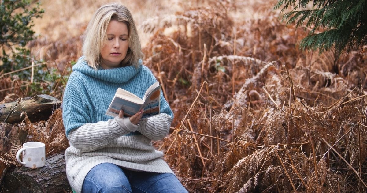 Sarah Robinson reading Wintering by Katherine May while sitting on a log in a quiet woodland.