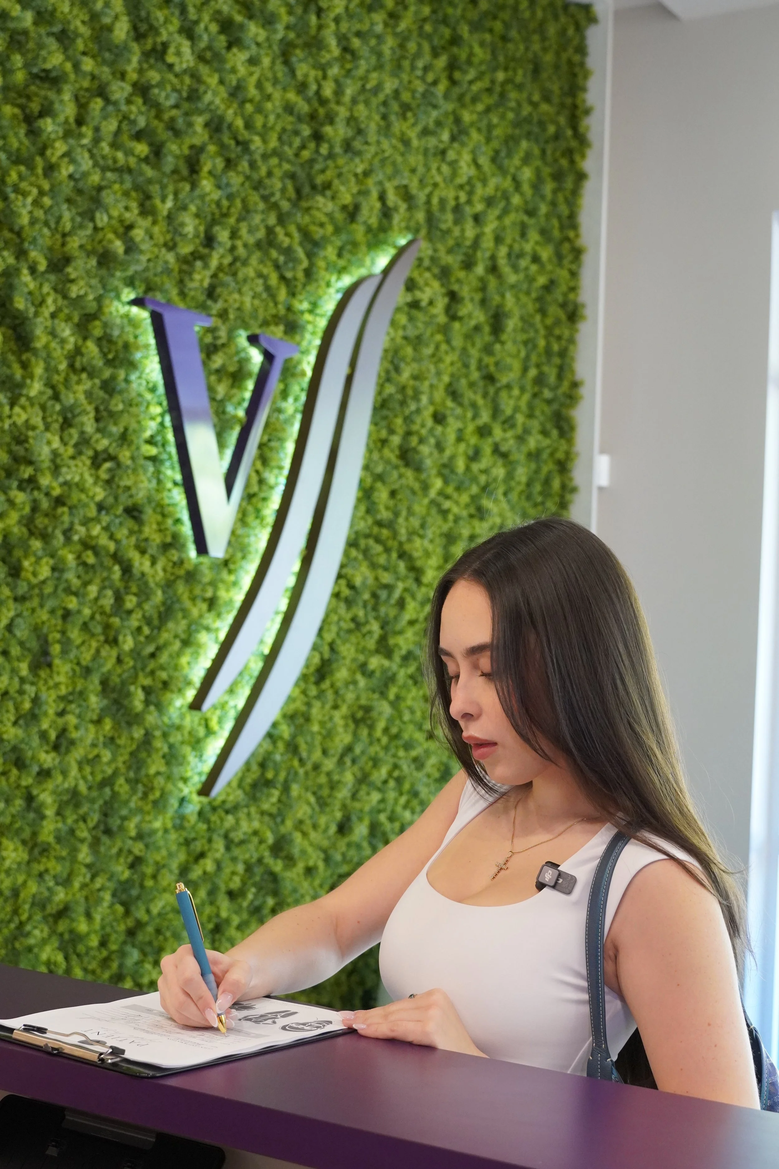 A young woman with long dark hair writing in a notebook at a reception counter, with a green leafy wall and a large illuminated logo behind her.