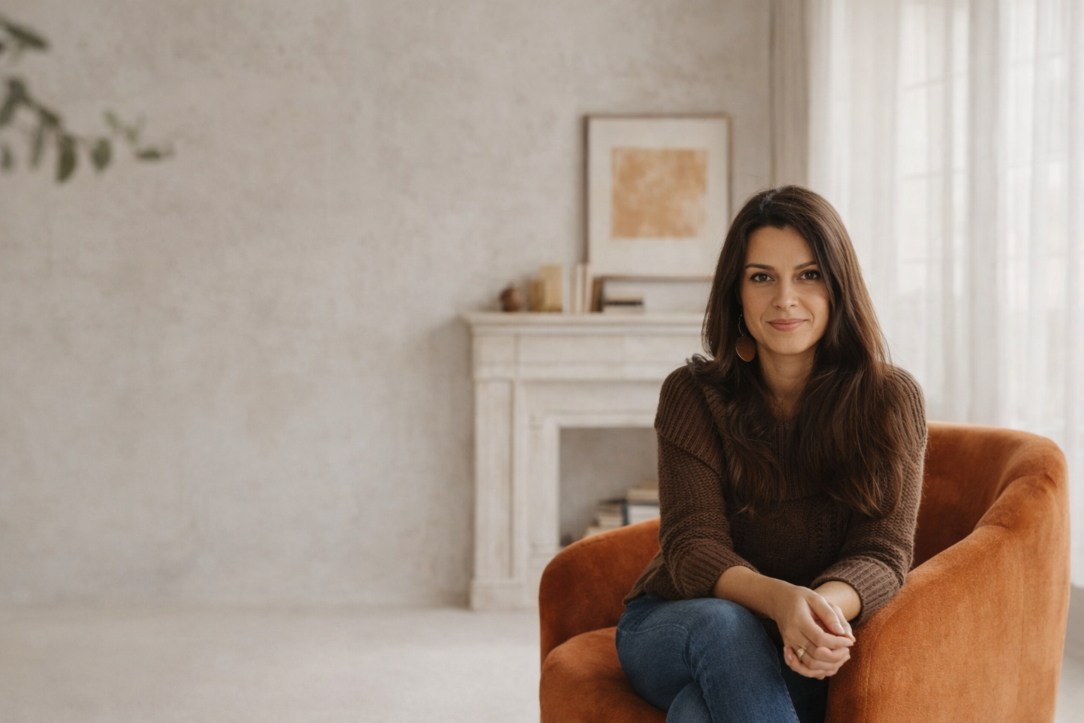 A woman with long brown hair and earrings, sitting on an orange chair in a cozy, well-lit room. She's smiling softly, wearing a brown sweater and blue jeans, with a white fireplace and bookshelf behind her.
