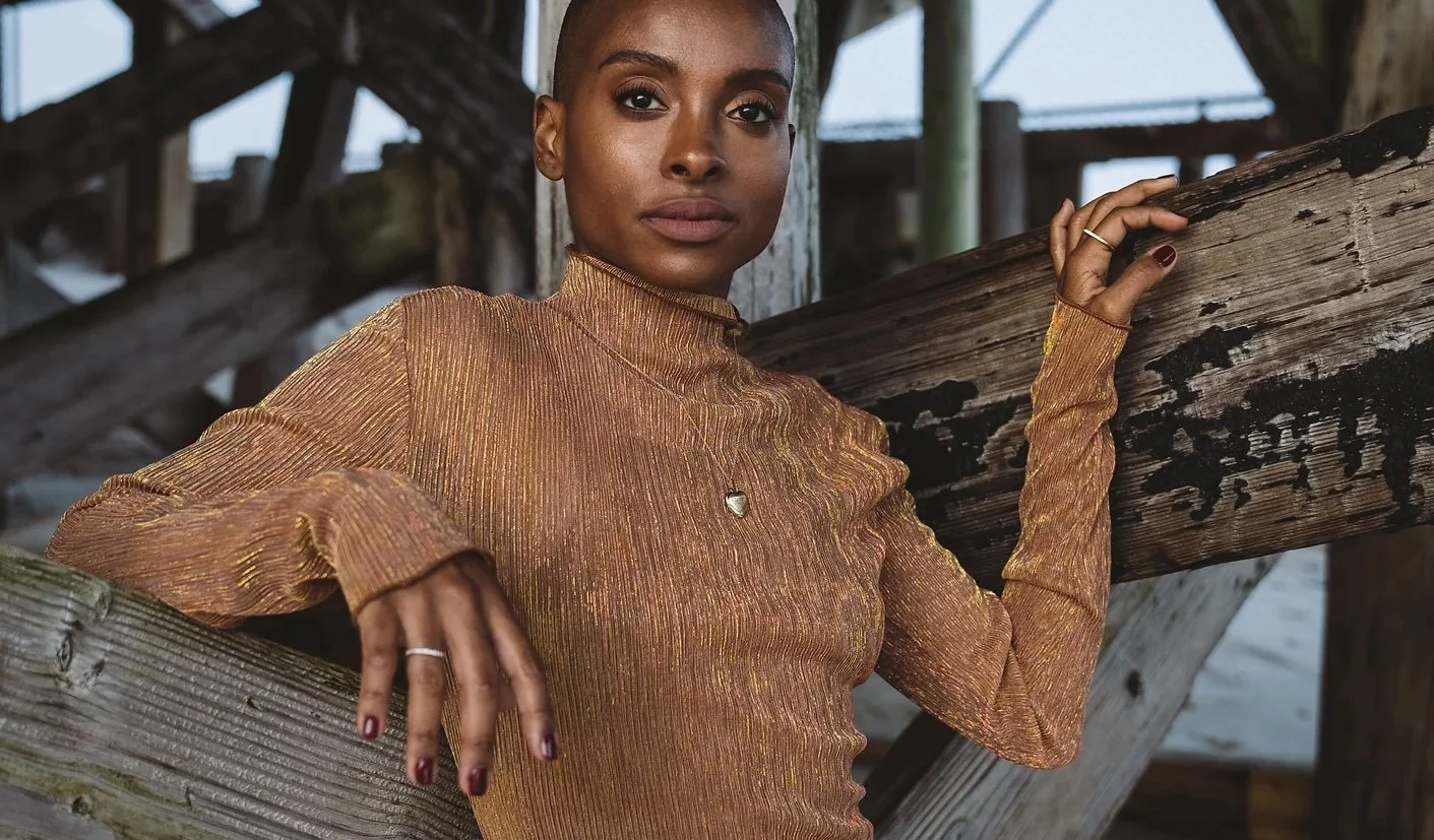 Person in a ribbed gold long-sleeve top leaning against wooden beams, outdoors.