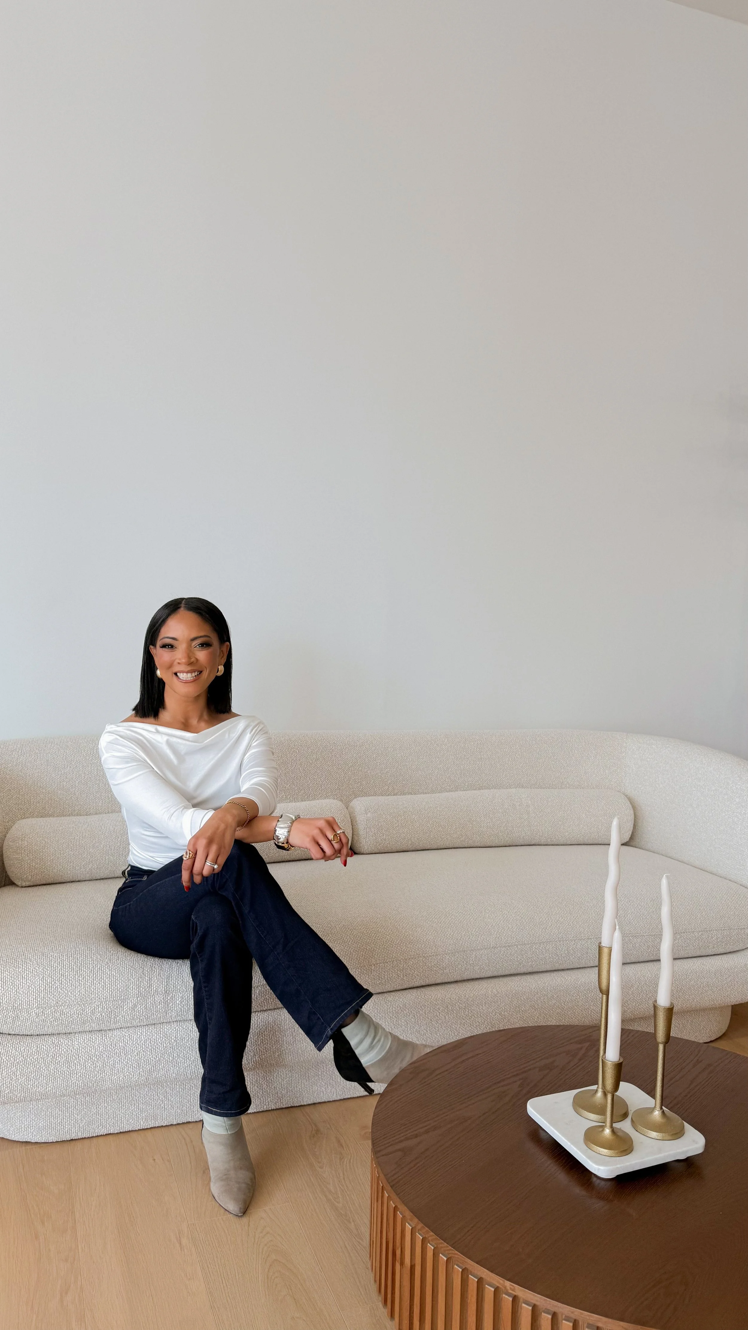 A woman sitting on a beige sofa, smiling, wearing a white top, dark jeans, and beige boots, with a plain white wall in the background and a wooden coffee table with decorative candles in brass holders.