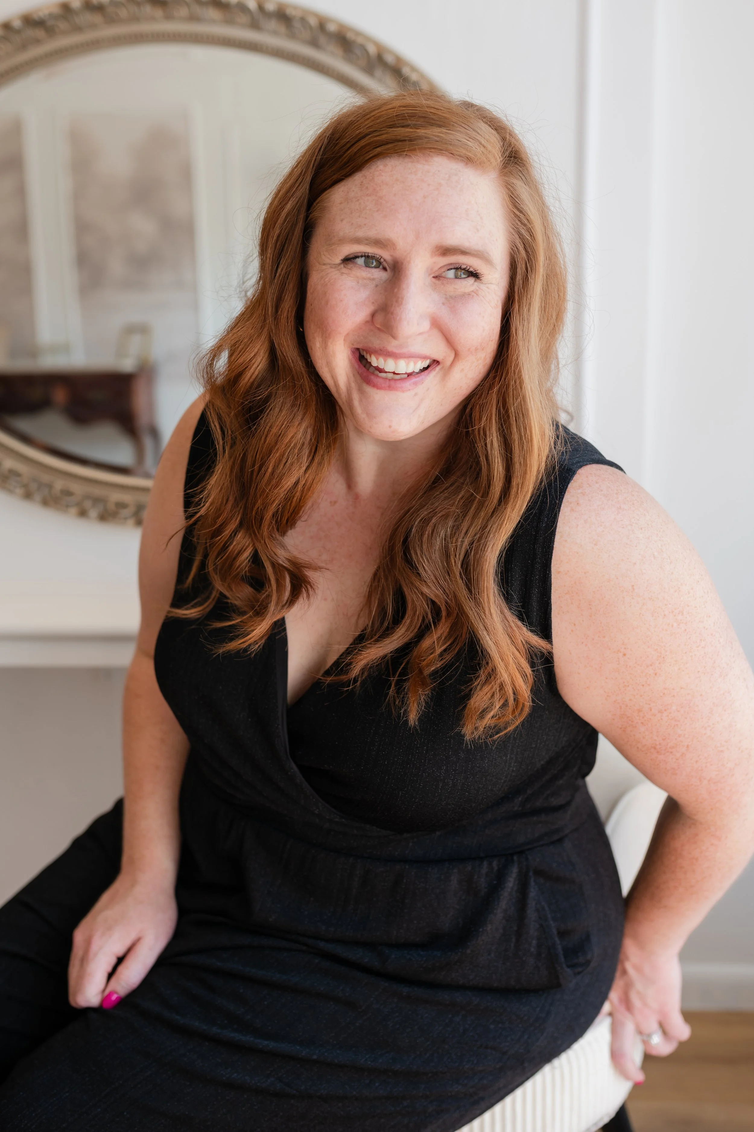 A woman with long red hair, freckles, wearing a black sleeveless dress, smiling and sitting in a room with a decorative mirror in the background.