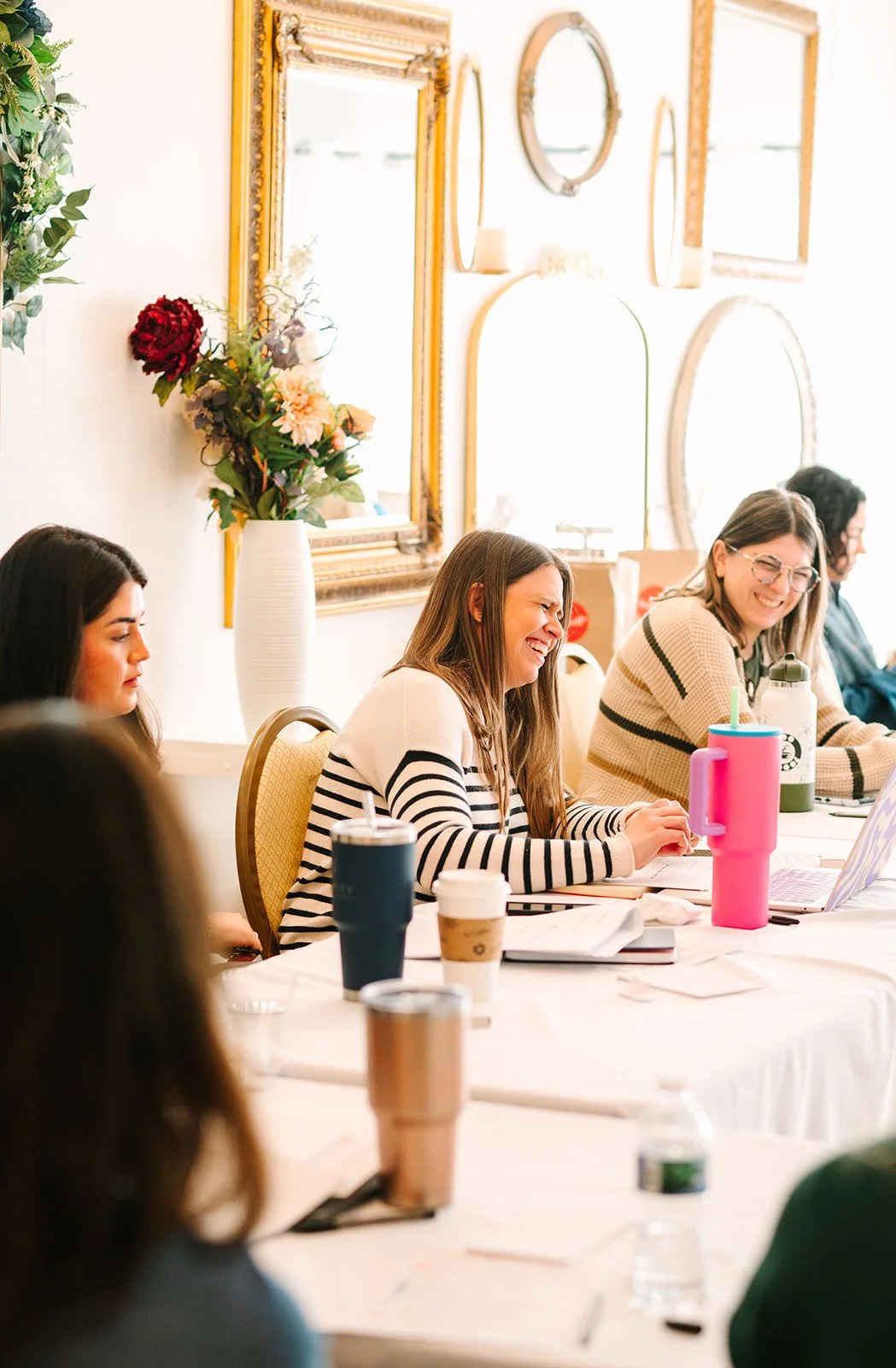 Women sitting at a long table, laughing and smiling during a meeting or workshop, with drinks and laptops in front of them, in a decorated room with mirrors and framed pictures on the wall.