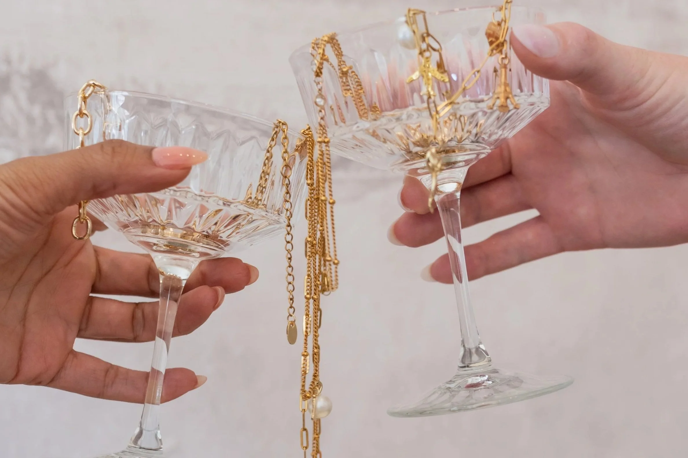 Two hands holding crystal cocktail glasses, with gold jewelry draped over the rims
