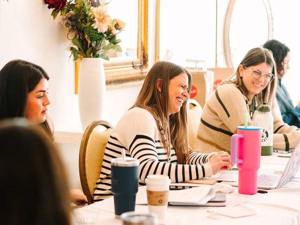 Group of women sitting at a table, smiling and laughing during a meeting or workshop in a bright room with a large mirror or window and a white flower vase in the background.
