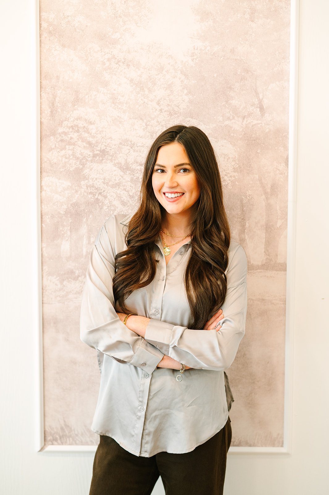 Smiling woman with long brown hair standing with arms crossed in front of a textured beige wall.