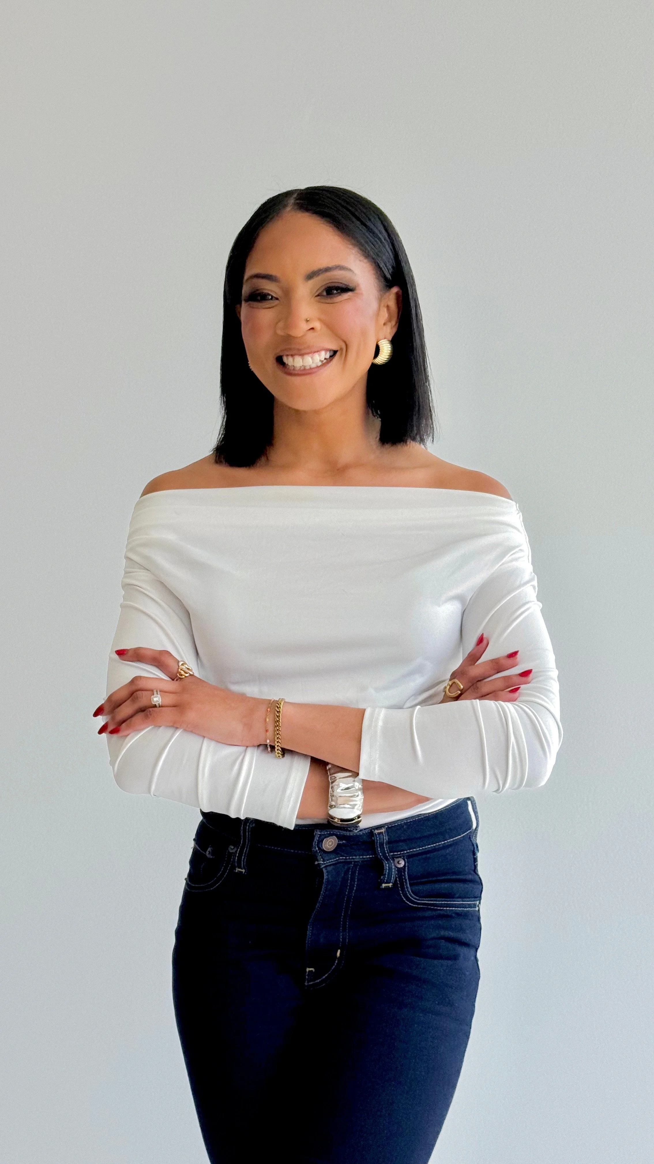 A woman with shoulder-length black hair, wearing a white off-shoulder top and dark jeans, standing with arms crossed, smiling against a plain light-colored background.