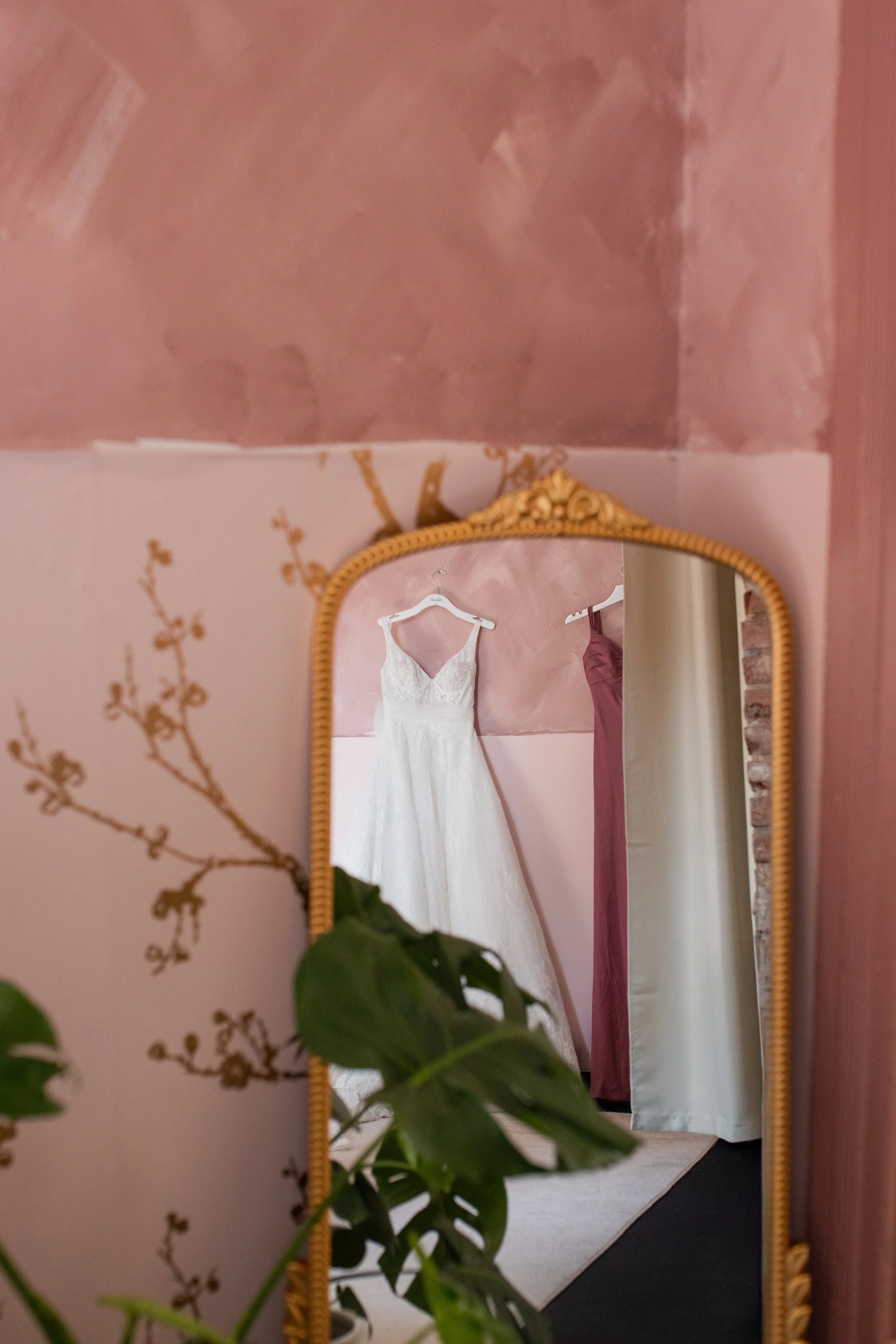 A wedding dress hanging on a white hanger in front of a mirror with a gold frame, reflecting a pinkest ceiling and a pink and white wall.