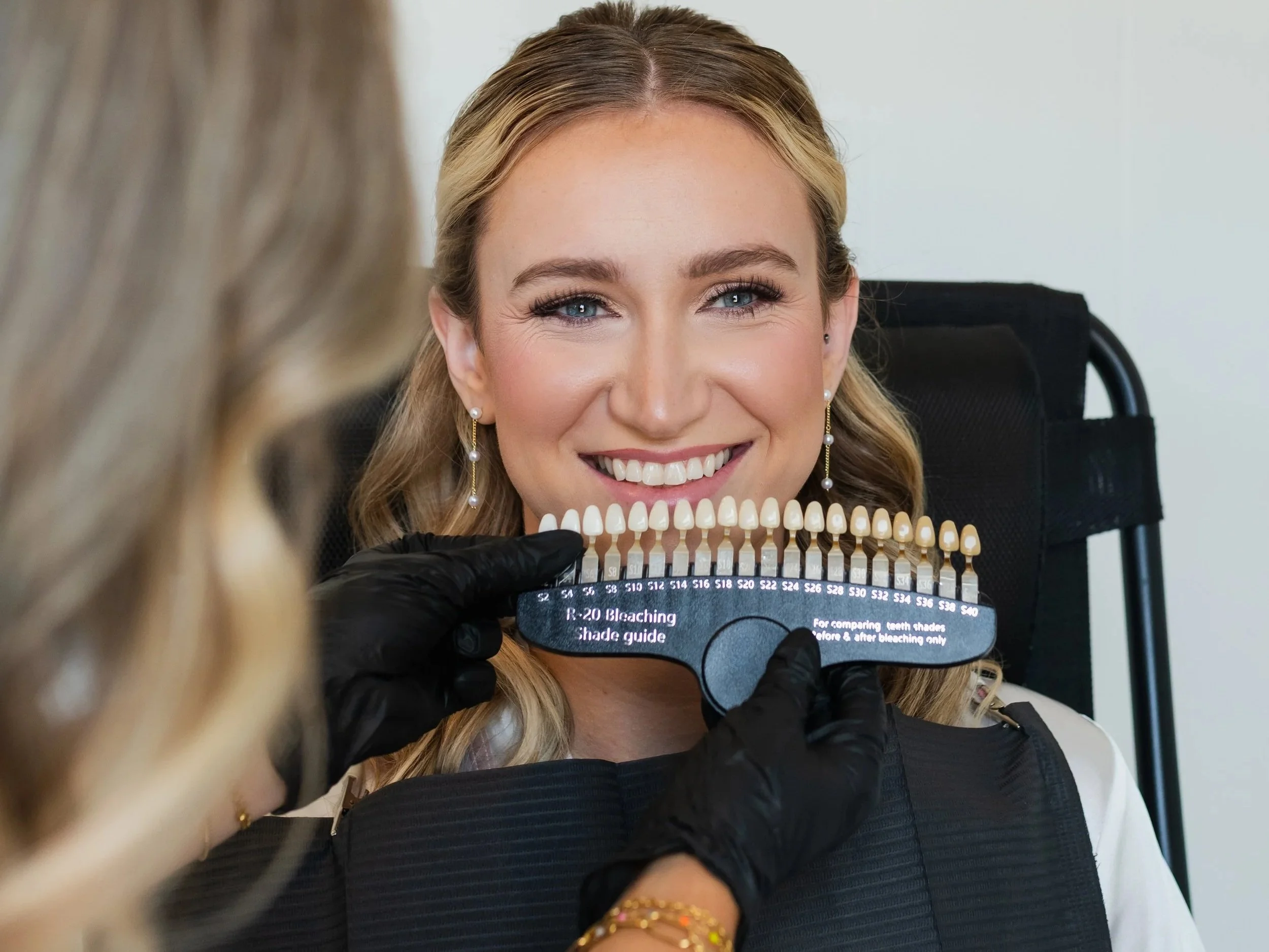 A woman with blonde hair sitting in a dental chair, smiling while a dental professional holds up a shade guide to her teeth to determine the best color for dental work.