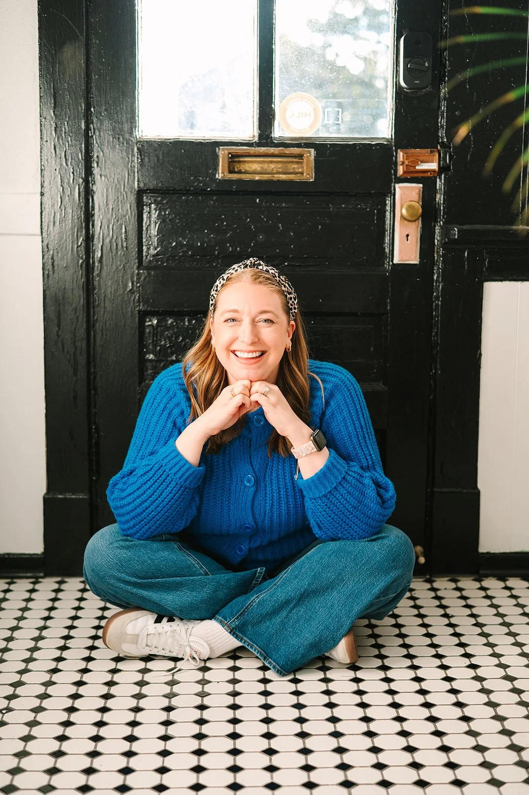 A woman sitting cross-legged on a black and white tiled floor, smiling, wearing a bright blue sweater, jeans, sneakers, and a leopard print headband, with a black door with a mail slot behind her.