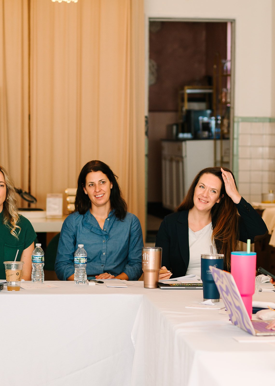 Women sitting at a conference table with water bottles and drinks, smiling and listening, in a room with beige panels and a kitchen in the background.