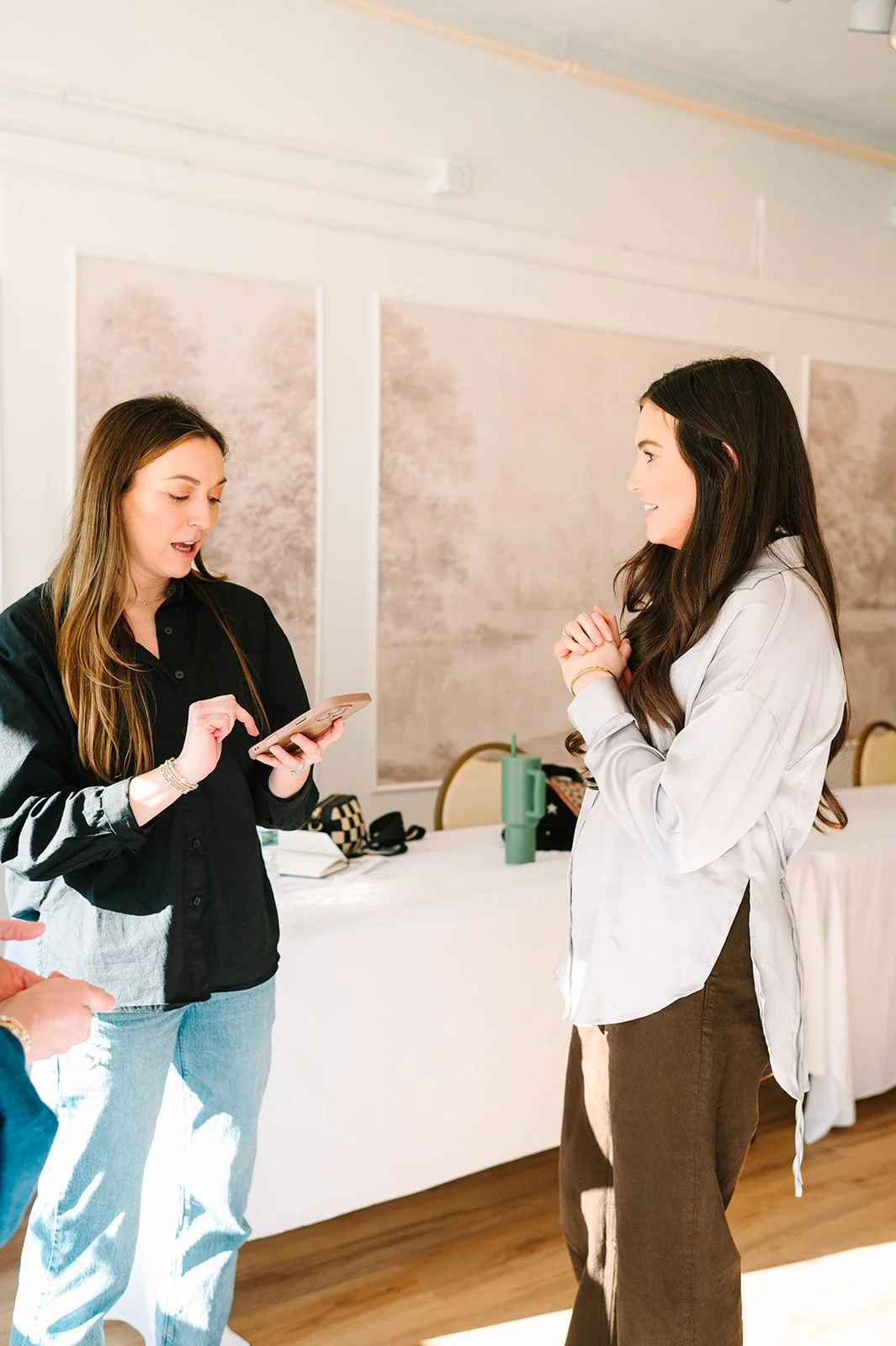 Two women are having a conversation in a well-lit room, one is holding a smartphone while the other listens.