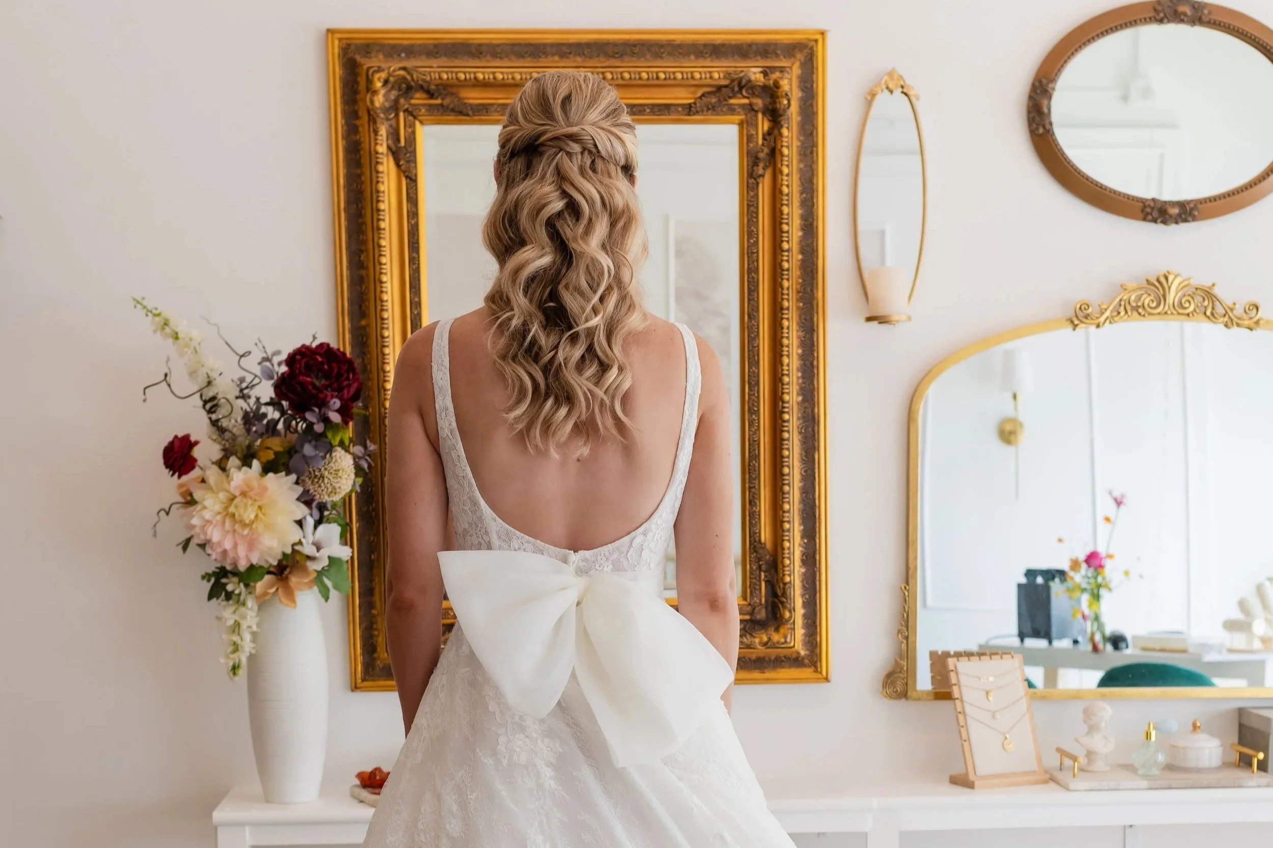 Back of a bride in a white wedding dress with a large bow on the back, standing in front of a decorative mirror with gold frame, surrounded by floral arrangements and decorative mirrors.