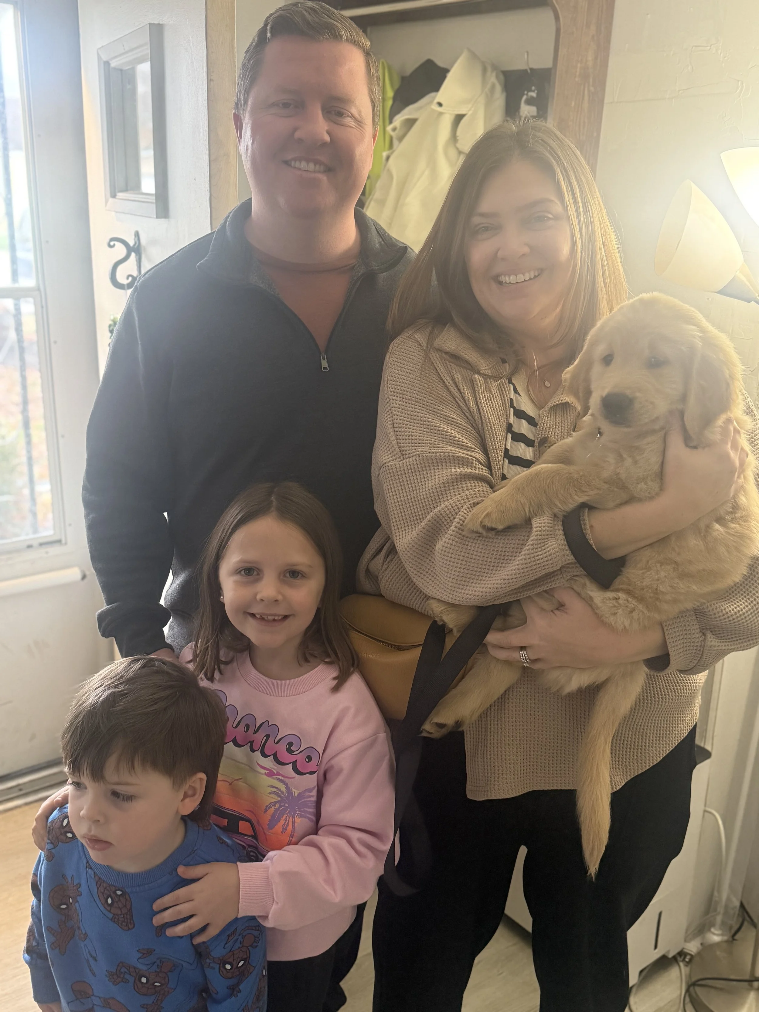 A smiling family of four with two children and two adults, one woman holding a golden retriever puppy inside a home.