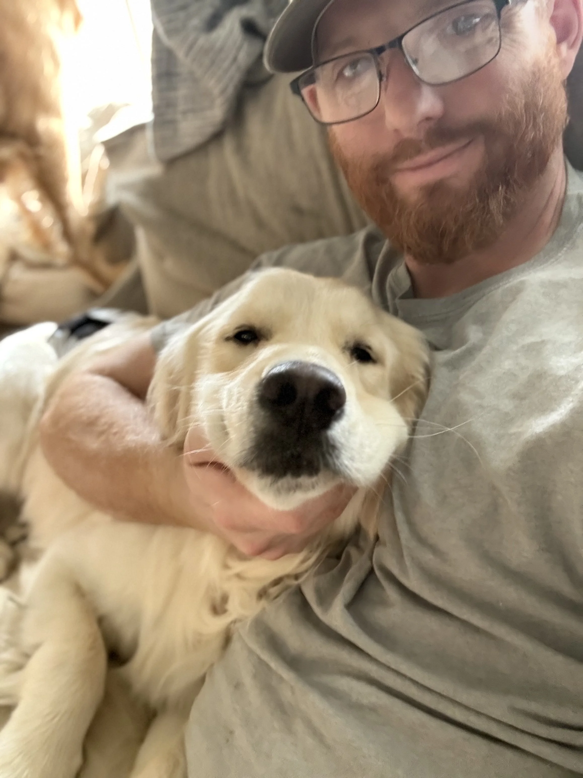 A man with glasses and a beard cuddling a golden retriever puppy on a sofa.