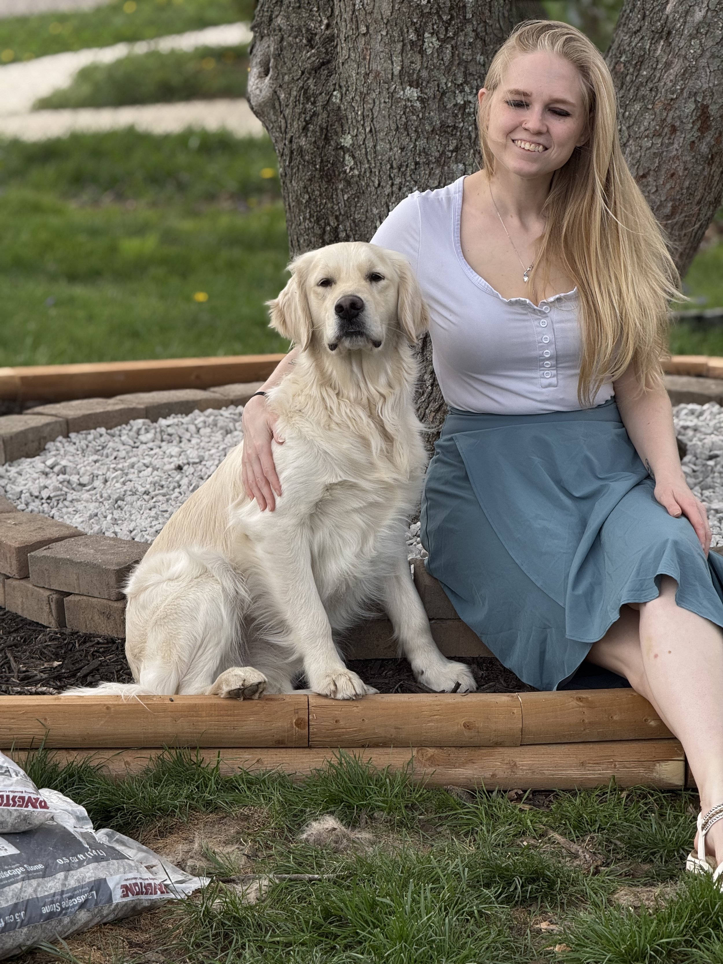 A woman with long blonde hair sitting on a garden border with a Golden Retriever dog, both sitting in front of a tree with green grass in the background.