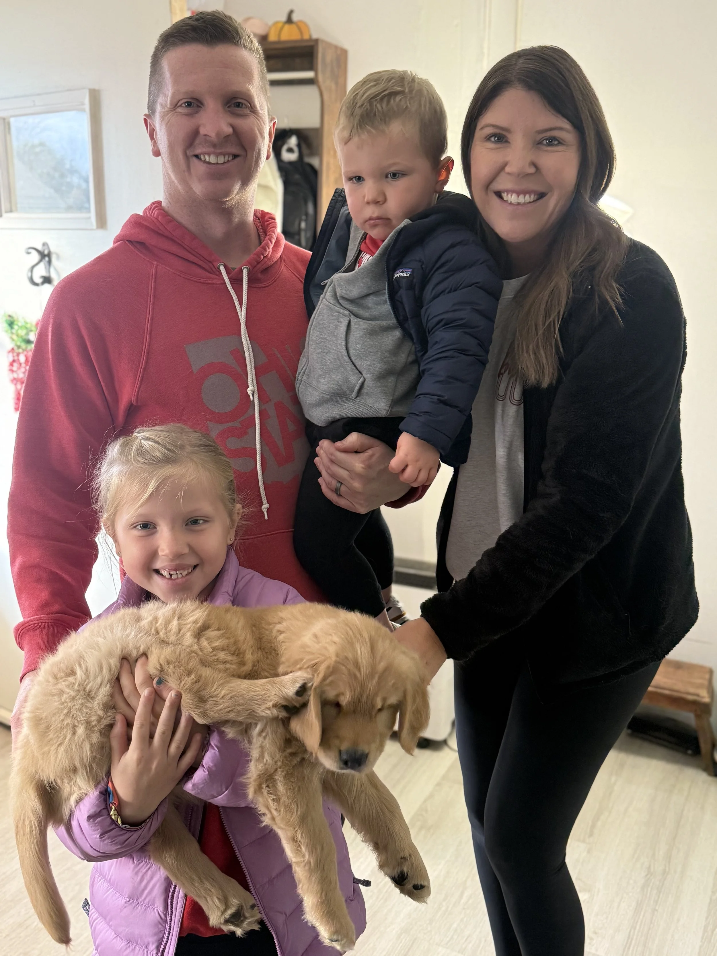 Family of four with a dog, including a man in a red hoodie, a woman with long brown hair, a young girl holding a golden retriever puppy, and a toddler in a jacket, standing in a cozy home interior.
