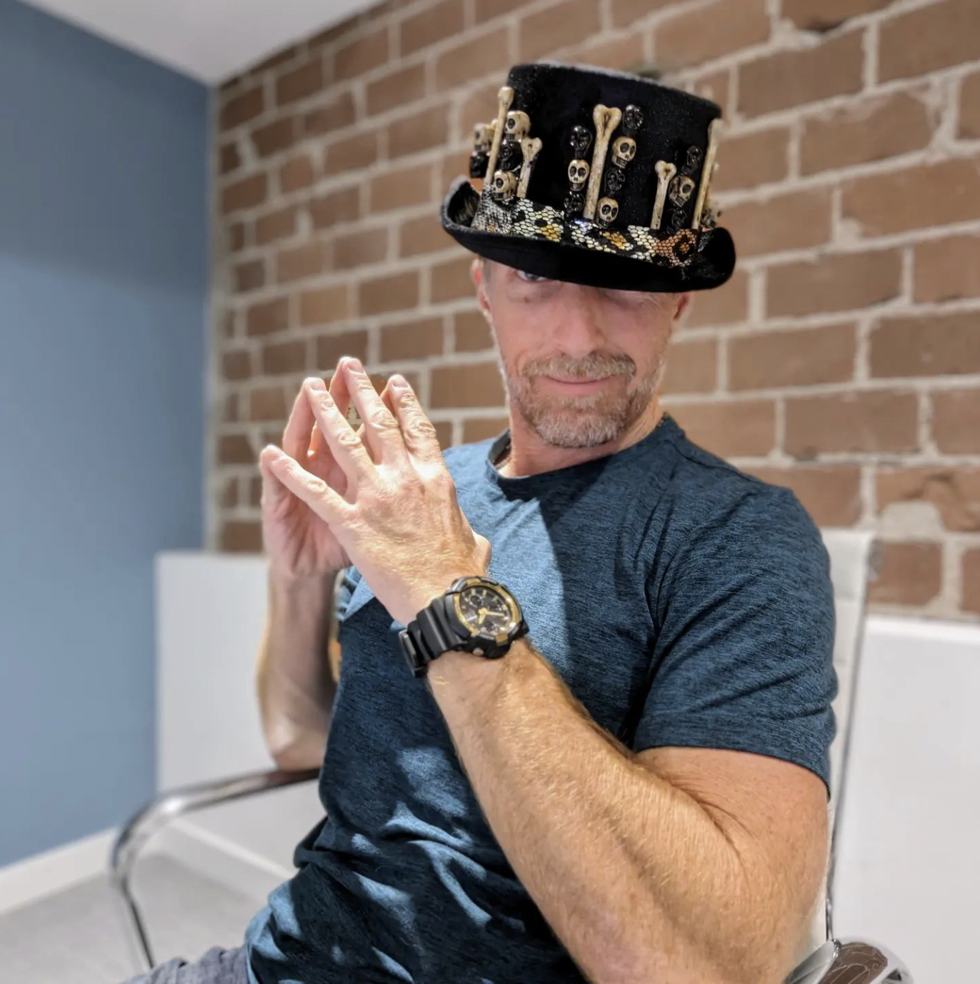 Man wearing a hat with skull and bone decorations, sitting in a chair, brick wall background.